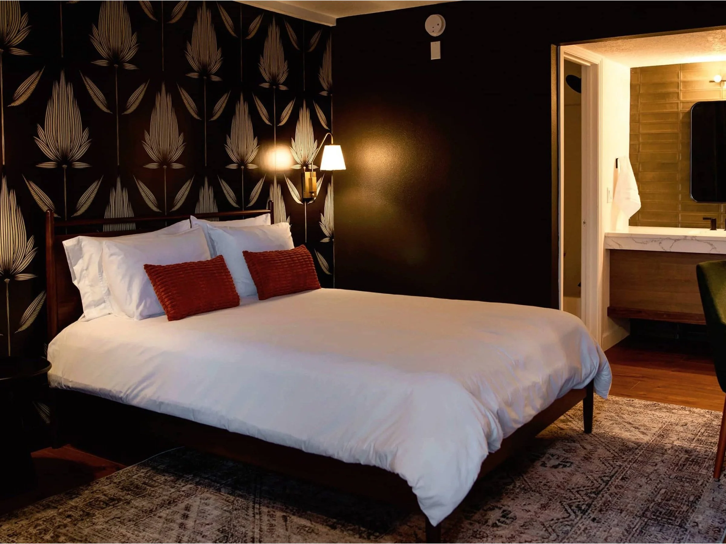 Bedroom with black patterned accent wall, white bed with white and red pillows, side wall with sconce, and a doorway leading to a bathroom with wooden walls and a white countertop.