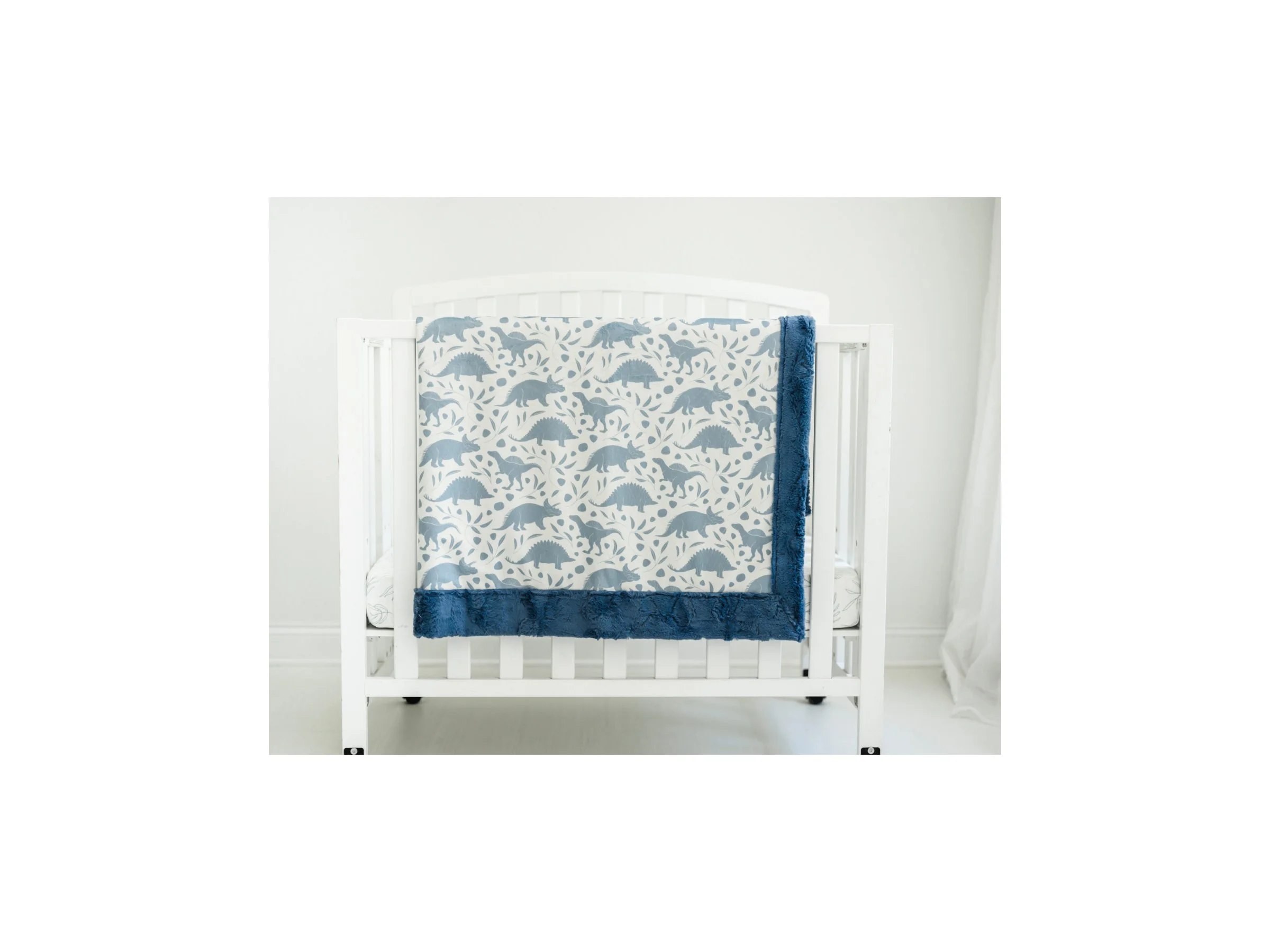 A white crib with a blue dinosaur print blanket hanging over the side and a blue blanket folded on top, set against a white wall and floor.