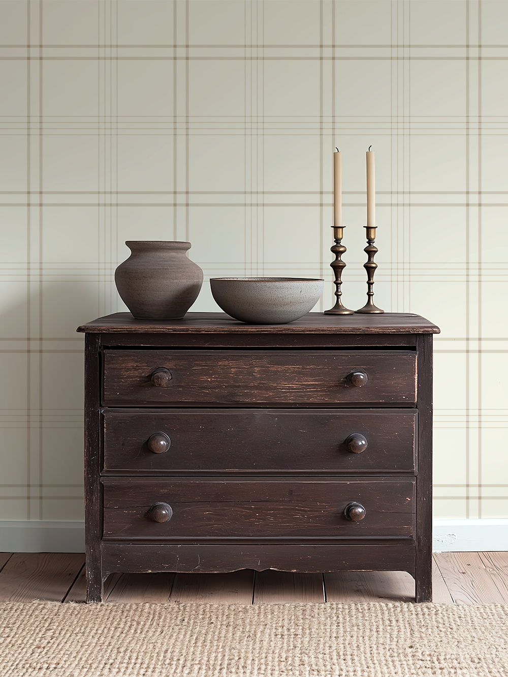 A dark wooden dresser with four drawers, decorated with two beige candles in holders, a ceramic vase, and a bowl, against a plaid wallpaper background.