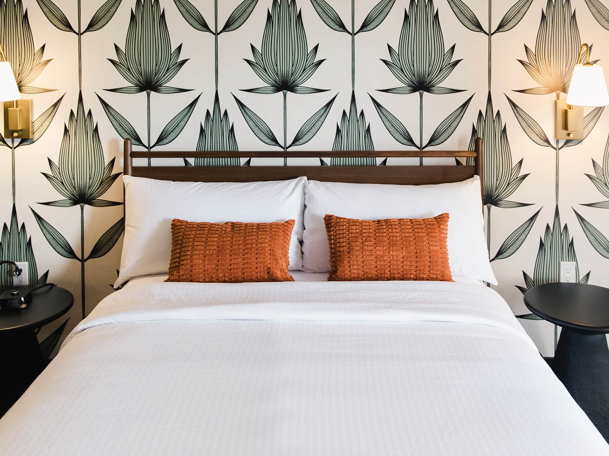A hotel room bed with white bedding and two orange textured pillows, set against a wall with black outlined large floral wallpaper, flanked by two wall-mounted lamps on either side.