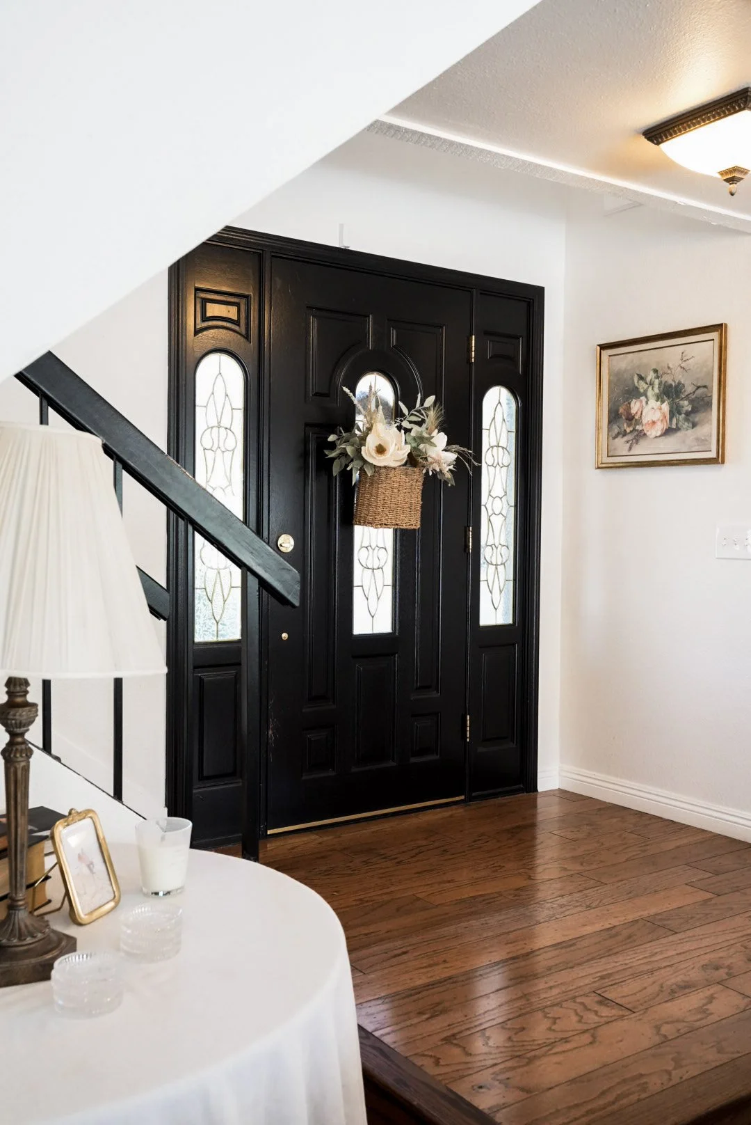 Close-up of a black front door with decorative glass panels, a floral wreath hanging on it, in a home entryway with hardwood floor, a wall painting, and a nearby table with a lamp, candles, and a picture frame.