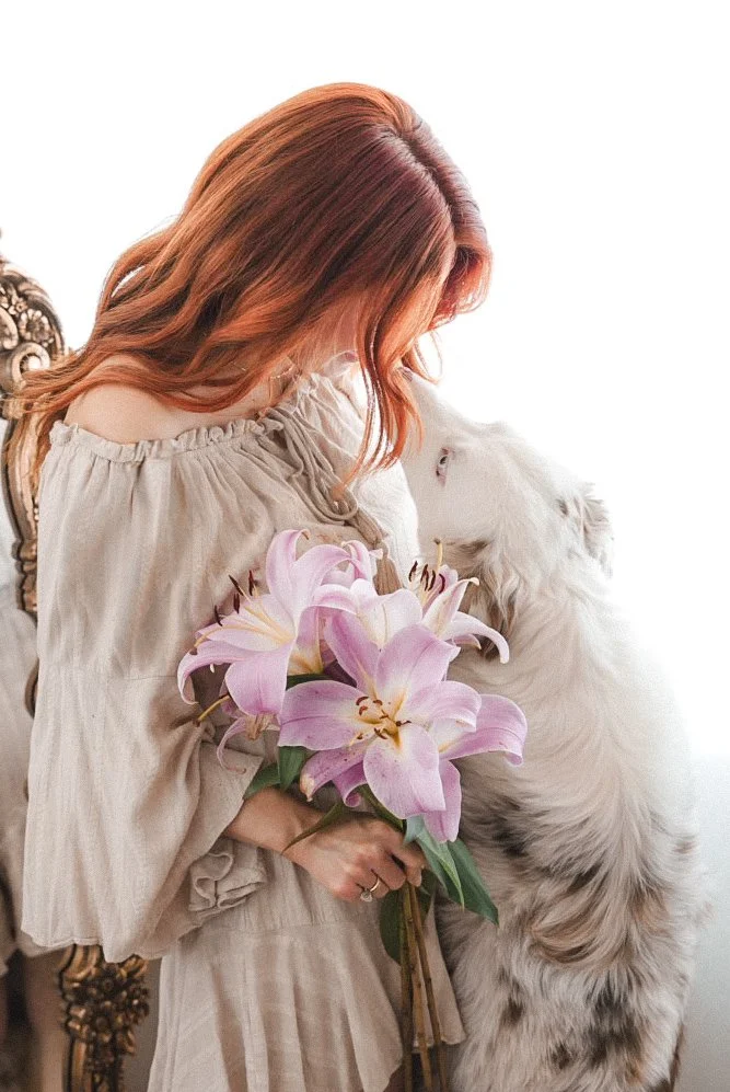 A woman with long, wavy red hair holding a bouquet of pink lilies, standing close to a white and gray dog.