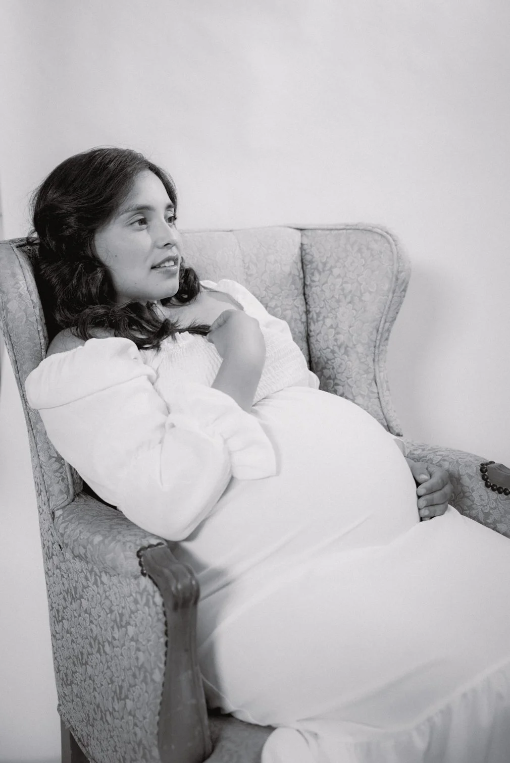 A pregnant woman relaxing on a patterned armchair, wearing a white dress, and gazing to her left with a calm expression.