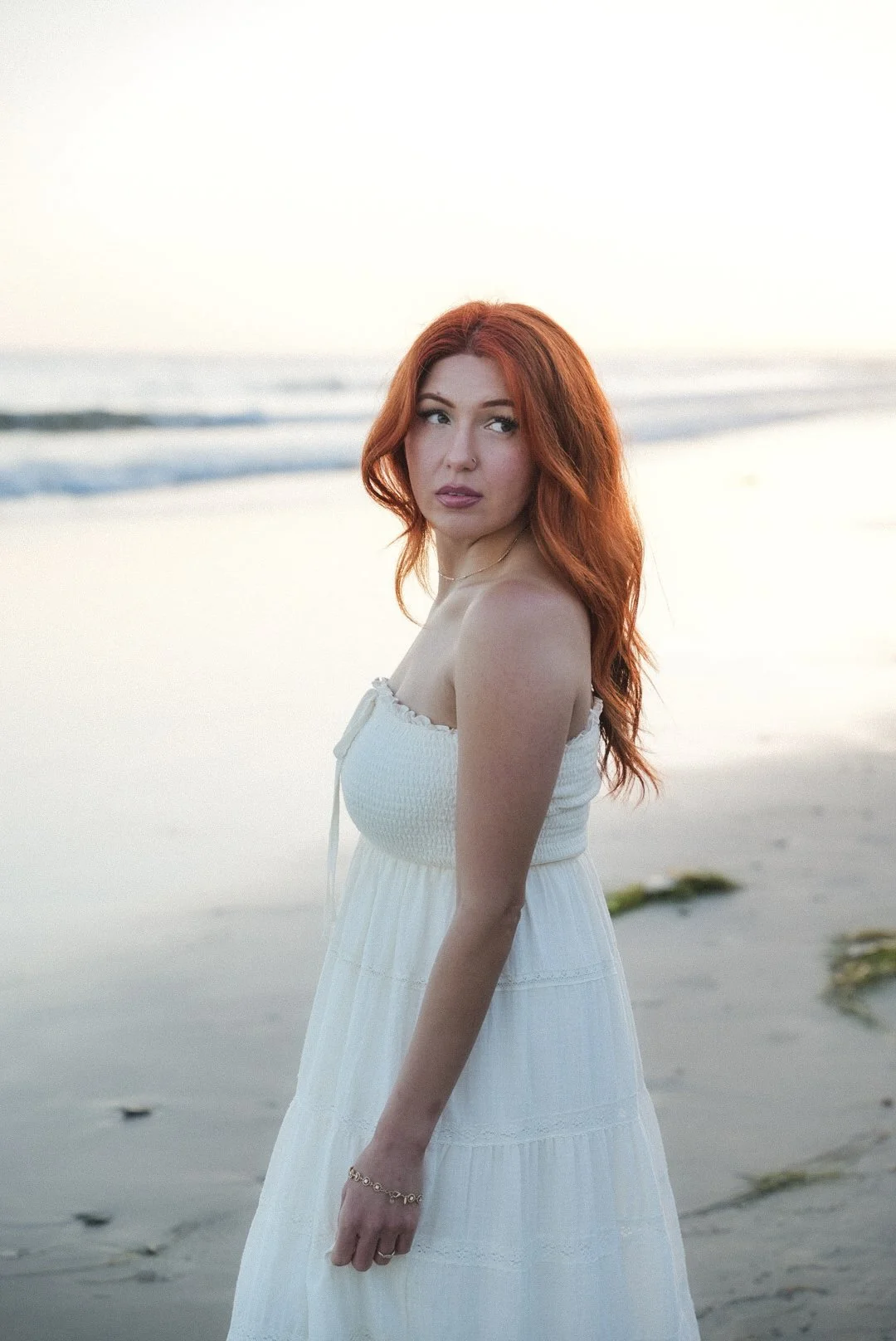A young woman with red hair in a white dress standing on a beach during sunset.