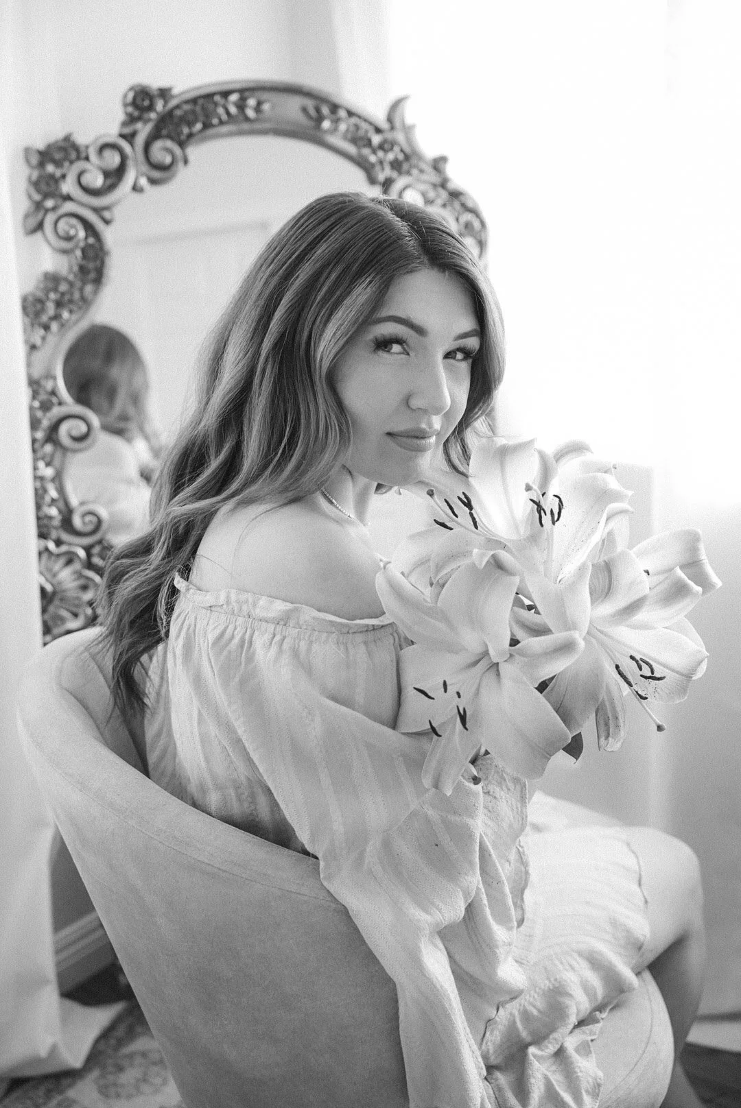 A woman with long wavy hair holding a large bouquet of lilies, sitting in a vintage armchair in front of an ornate mirror. The setting is elegant and softly lit, with a mirror reflecting part of her back.