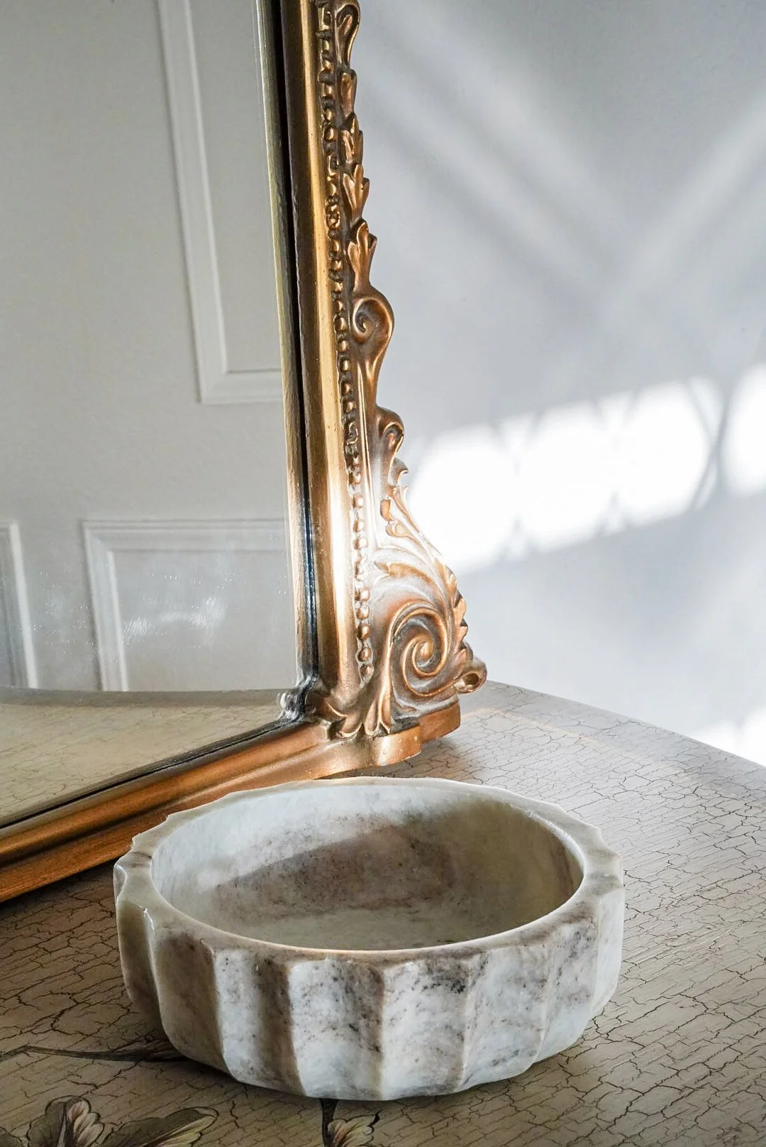 A vintage wooden framed mirror, a ceramic bowl with a scalloped edge, and a table with a cracked, textured surface are arranged together. The mirror reflects a light-colored wall with decorative molding.