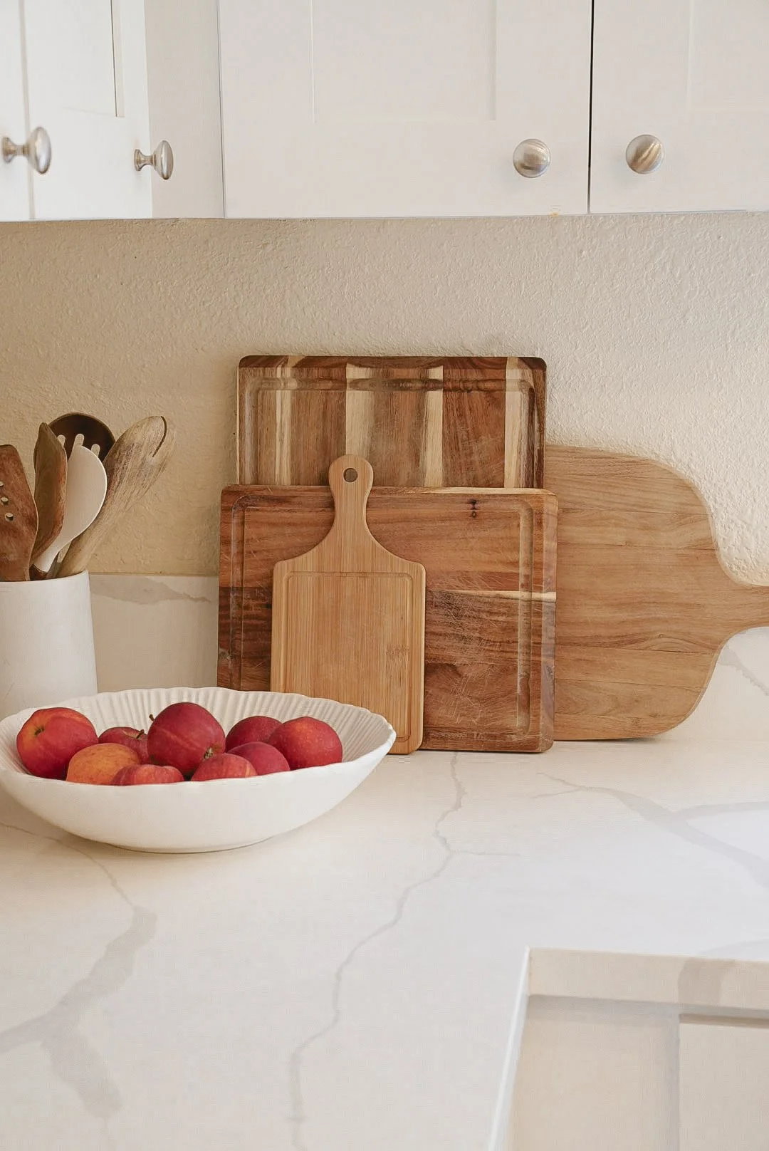 White bowl of red apples on a white kitchen countertop with wooden cutting boards and utensils in the background.
