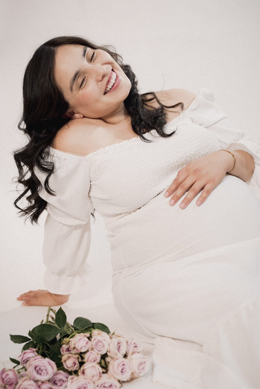 A woman with long dark hair smiling and sitting on a white surface, surrounded by pink roses, wearing a white off-shoulder dress, with a relaxed and joyful expression.