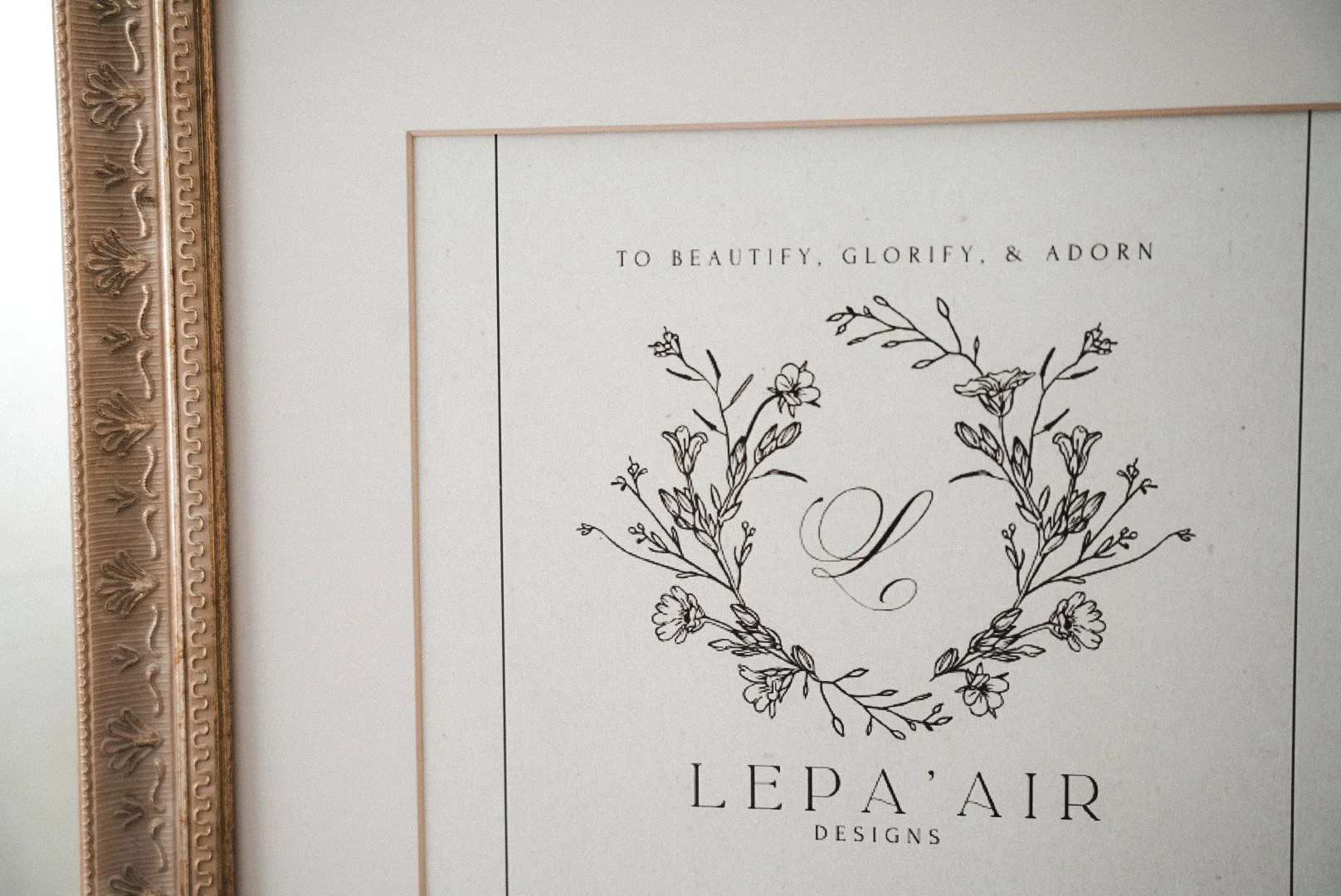 Decorative framed sign with floral design and text that reads "To beautify, glorify, & adorn" with a large cursive letter 'L' and the words "Lepa Air Designs" below.