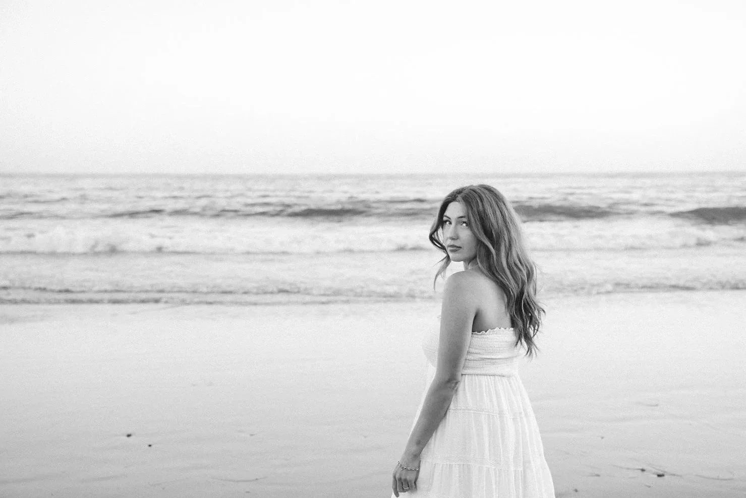 A woman in a white dress standing on a beach with the ocean in the background, looking over her shoulder.