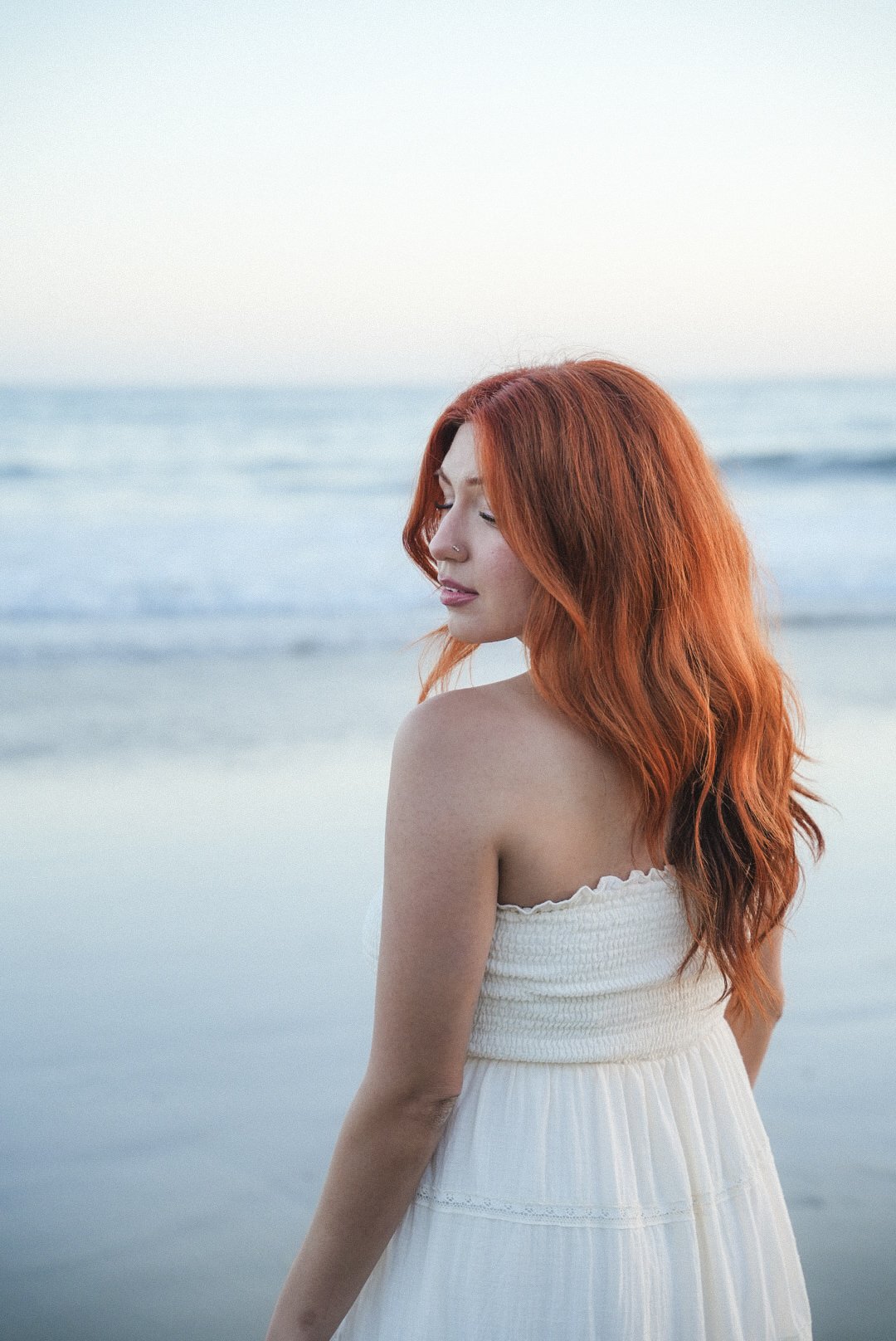 A woman with long red hair standing on a beach with calm waves in the background, wearing a strapless white dress with her eyes closed.