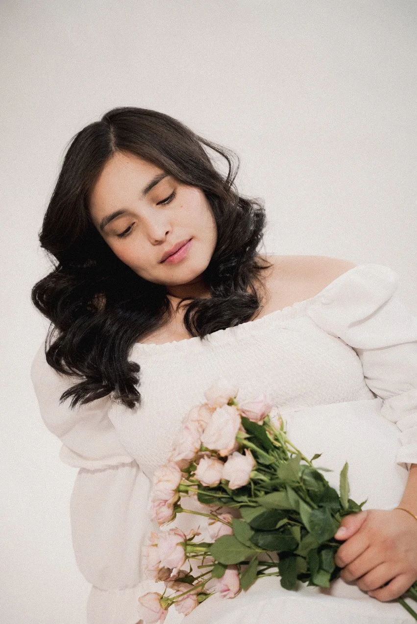 A woman with dark, wavy hair and closed eyes holding a bouquet of pink roses, wearing an off-shoulder white dress, standing against a plain light-colored background.