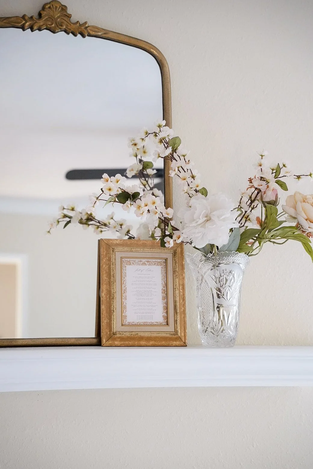 A decorative arrangement of white and pink flowers in a glass vase, a gold-framed document, and a large ornate mirror on a white shelf against a beige wall.