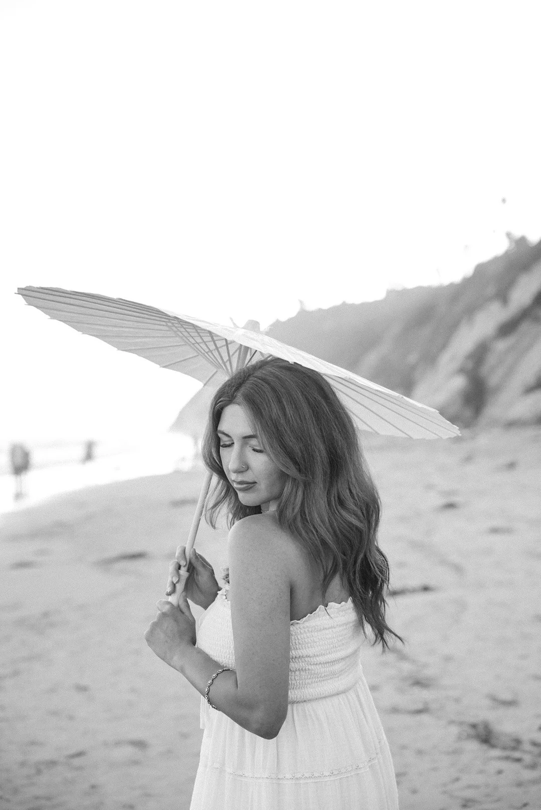 Black and white photo of a young woman with wavy hair holding a parasol at the beach, standing with her eyes closed and a serene expression.