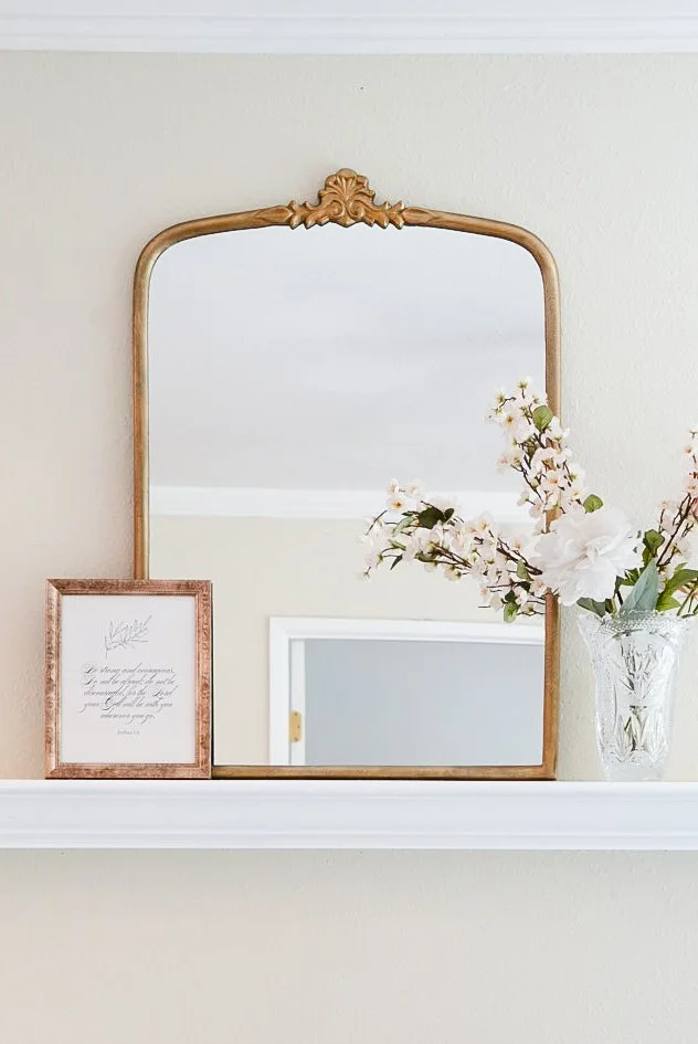 Decorative wall mirror with gold frame, a framed quote, and a vase with white and pink flowers on a white shelf.