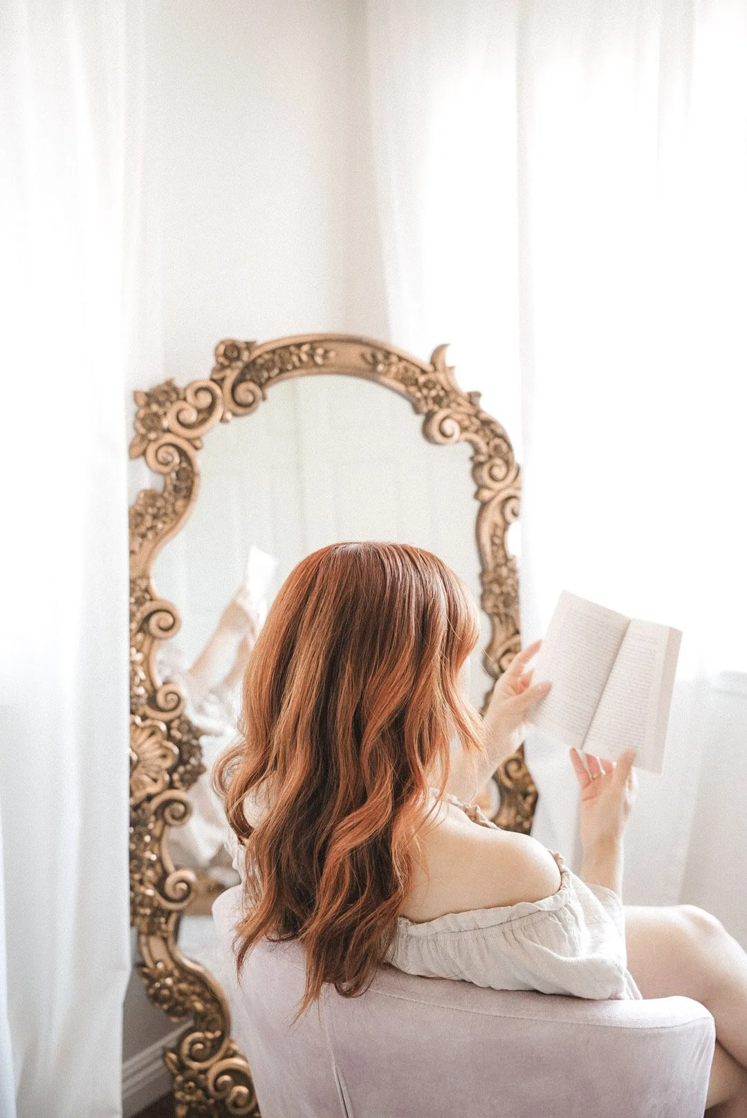 A woman with long, wavy red hair sitting in a pink armchair, reading a book, with a large ornate mirror behind her reflecting her reading.