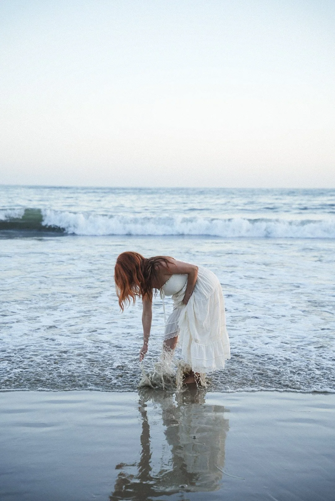 A woman with red hair in a white dress standing in the shallow surf at the beach, bending over to touch the water.