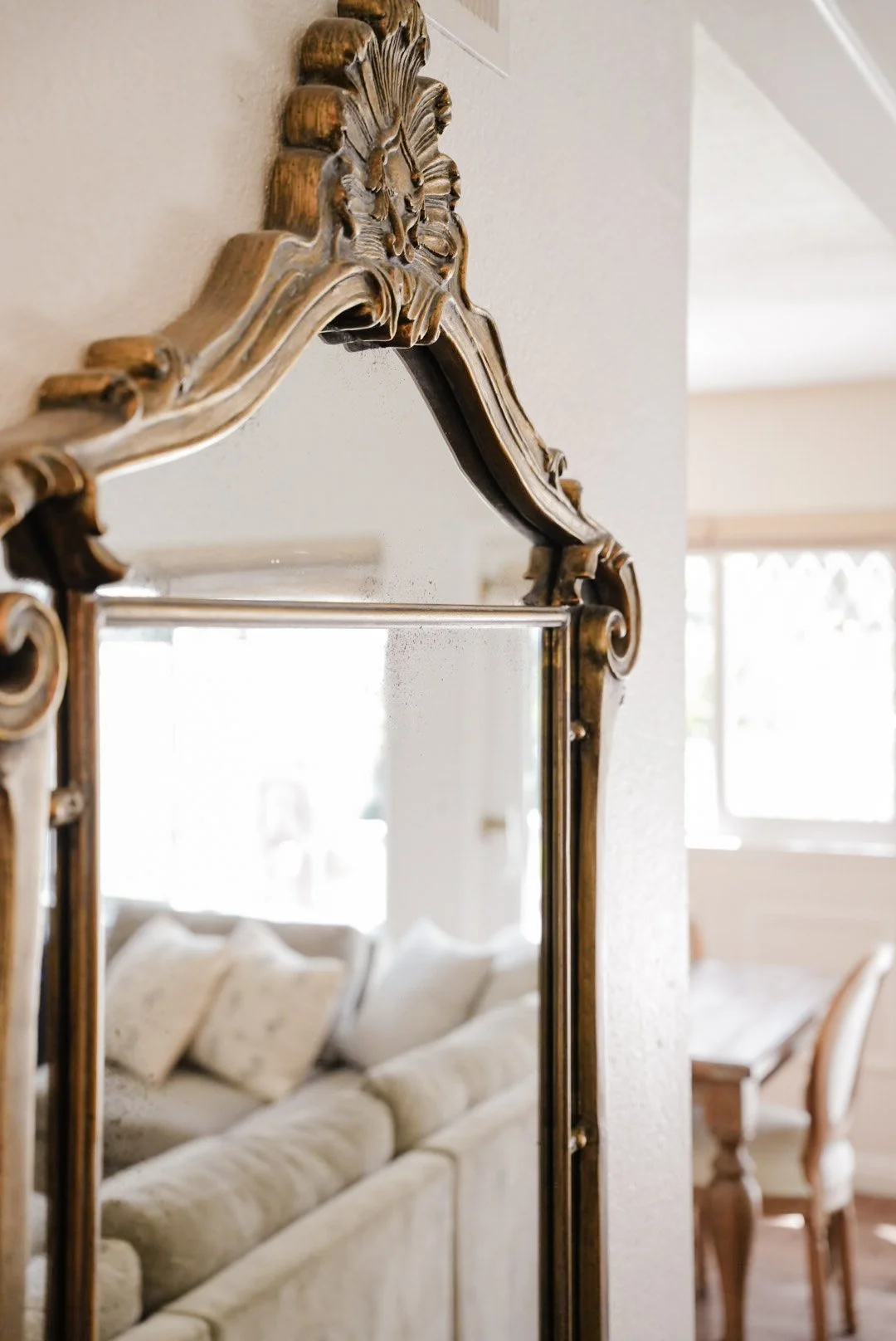 Close-up of an ornate, vintage-style mirror with a carved wooden frame hanging on a wall, reflecting a white sofa with pillows and part of a living room with windows and chairs.