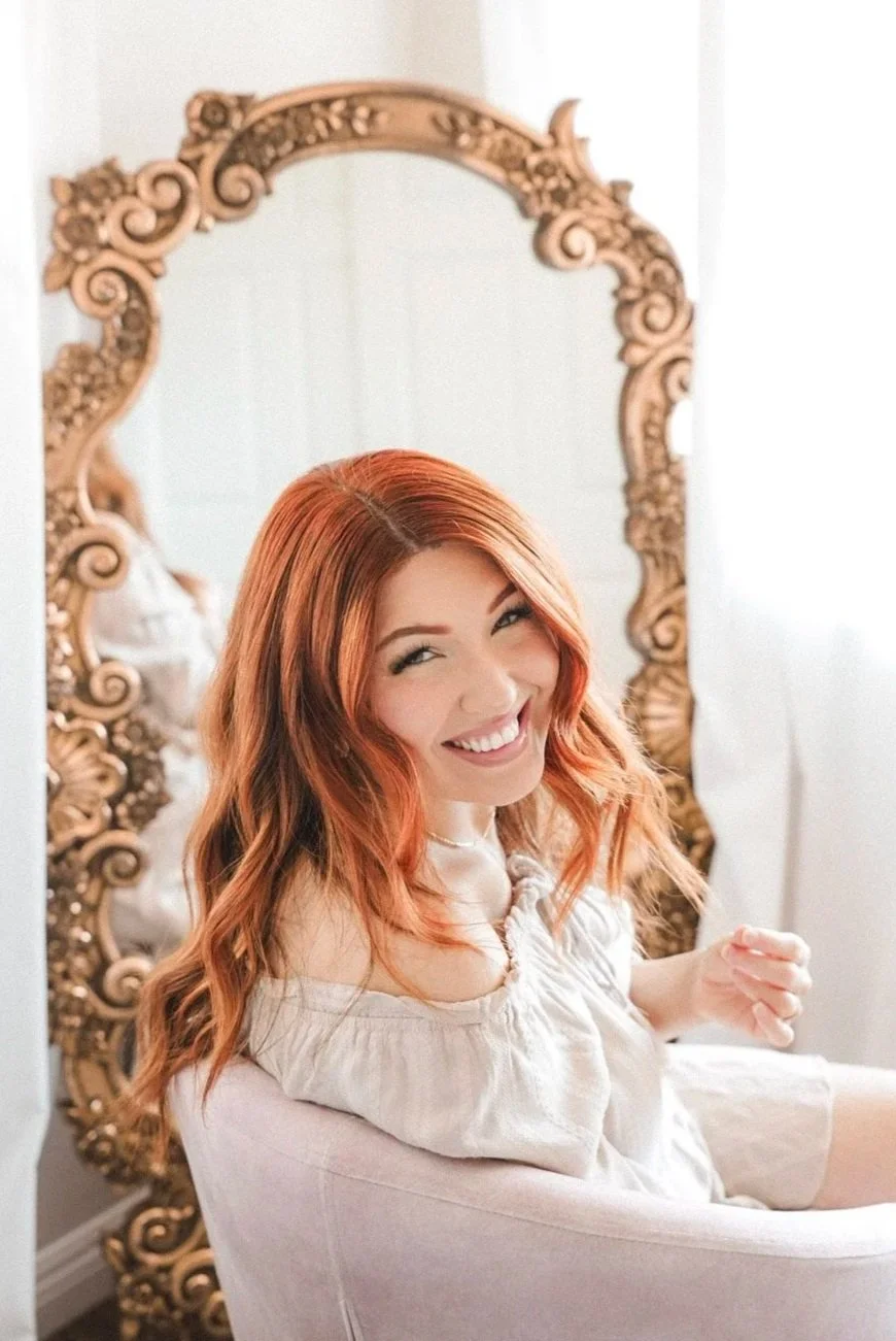 A woman with red hair smiling while sitting in front of an ornate gold mirror.