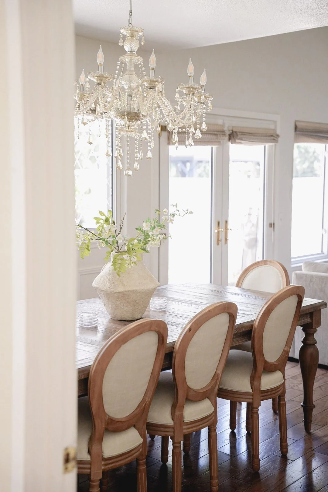 A dining room with a wooden table, five cream upholstered chairs, a chandelier hanging from the ceiling, a large vase with white flowers and greenery, and three large windows with beige blinds.