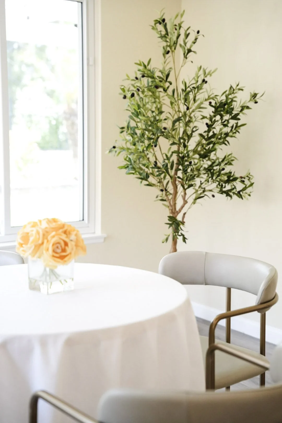 A round dining table with a white tablecloth, decorated with a bouquet of peach-colored roses in a square glass vase. Behind the table, there are white and wood chairs, a large window on the left, and an indoor green plant in the corner.