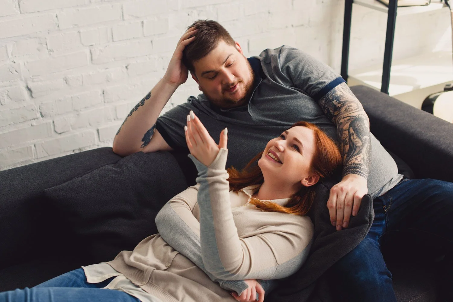 A young man with tattoos and a woman with red hair sitting on a black sofa, smiling and laughing while talking and making eye contact in a cozy indoor setting.