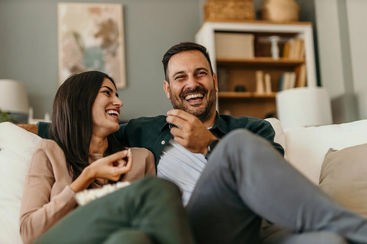 A couple sitting on a white couch, laughing and enjoying each other's company, with a bookshelf and framed art in the background.