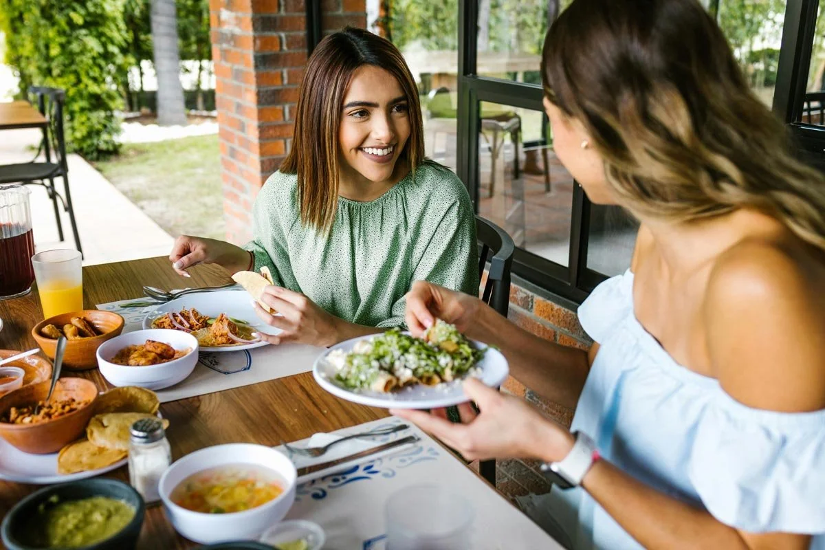 Two women sharing a meal at a restaurant table outside, with various dishes including salads, bowls, and drinks.