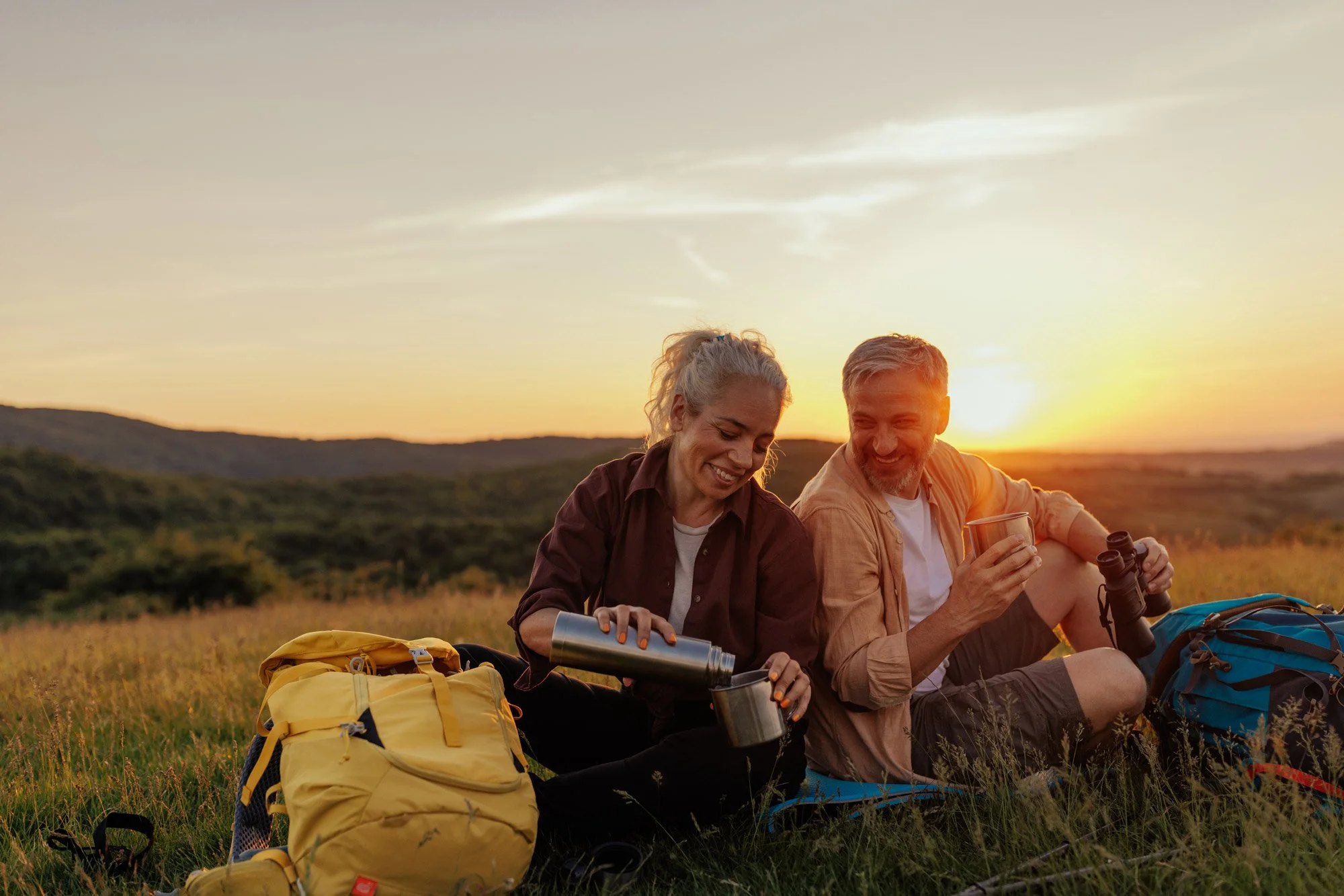 Two smiling people, a woman and a man, sitting on the grass during sunset, preparing for a hike with backpacks, water bottles, and binoculars around them.