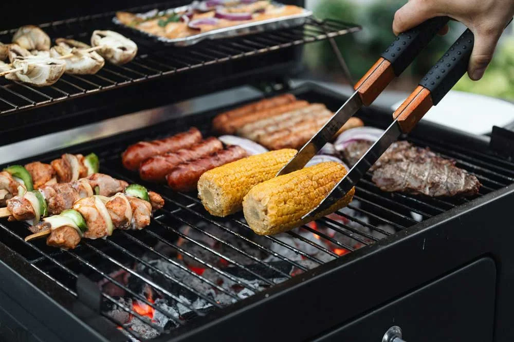 Person using tongs to grill corn on the cob, sausages, steak, chicken skewers, and hot dogs on a barbecue grill outdoors.
