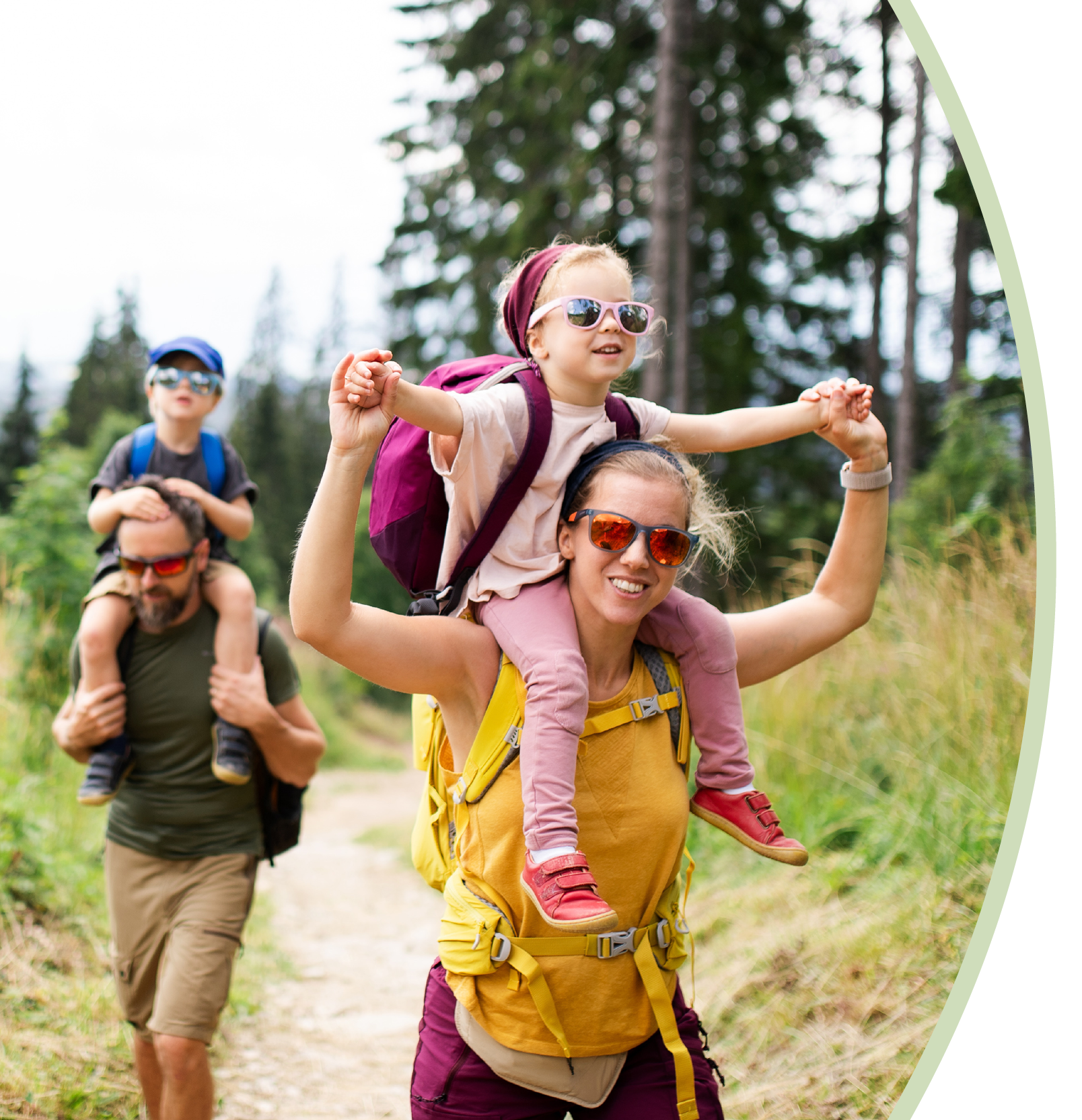 A family hiking outdoors together in a forest: a woman carrying a young girl on her shoulders with sunglasses, a man walking with a backpack behind them.