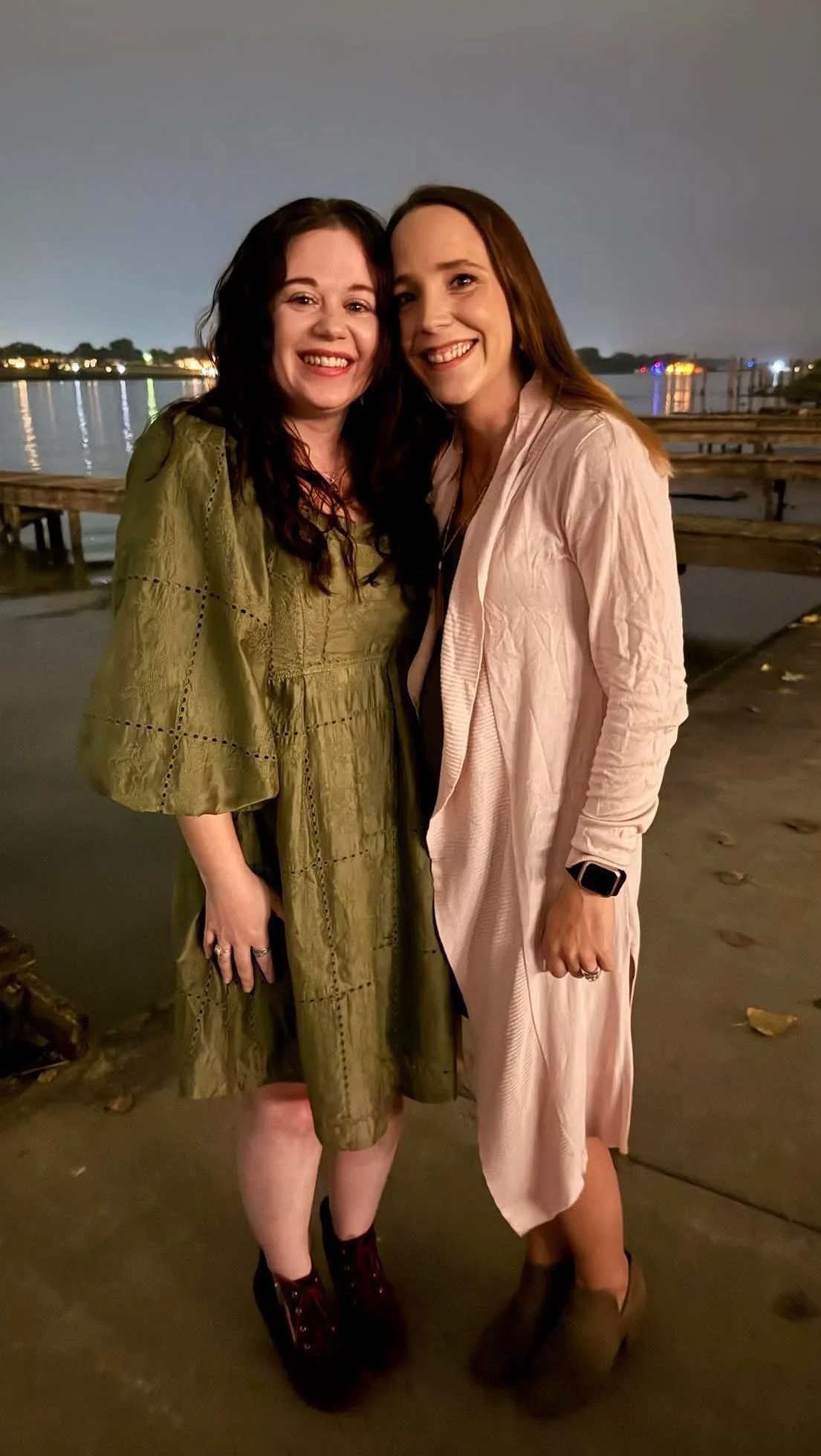 Two women smiling and posing together outdoors at night, near a body of water with reflections of lights, standing on a dock or pier.