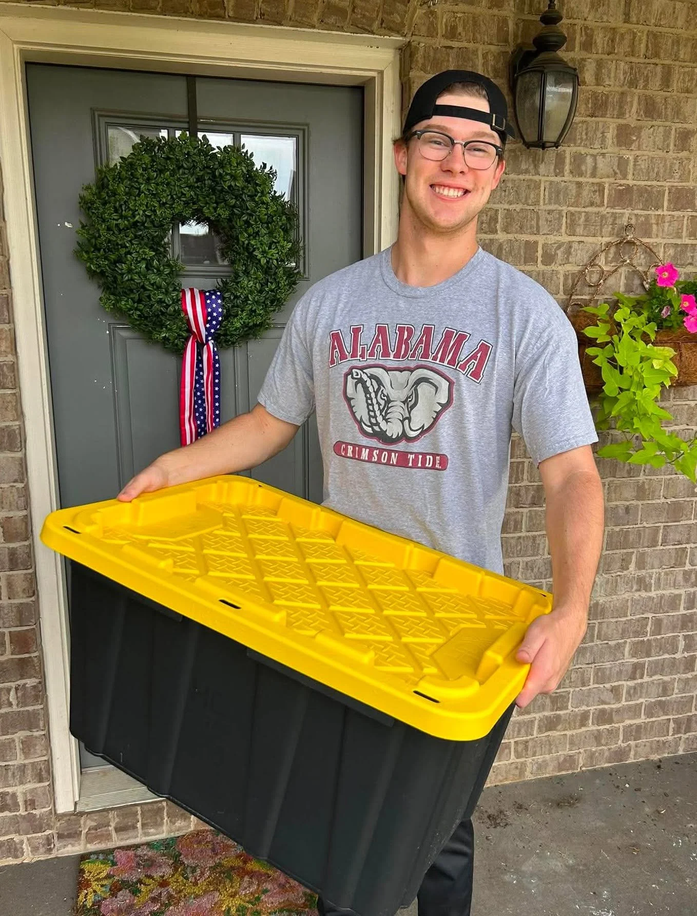 A young man wearing glasses, a black baseball cap worn backwards, and a gray Alabama Crimson Tide T-shirt is smiling while holding a large black and yellow plastic storage container in front of a brick house door. There is a festive green wreath with a red, white, and blue striped ribbon hanging on the door and a hanging basket with pink flowers and green foliage on the wall to his right.