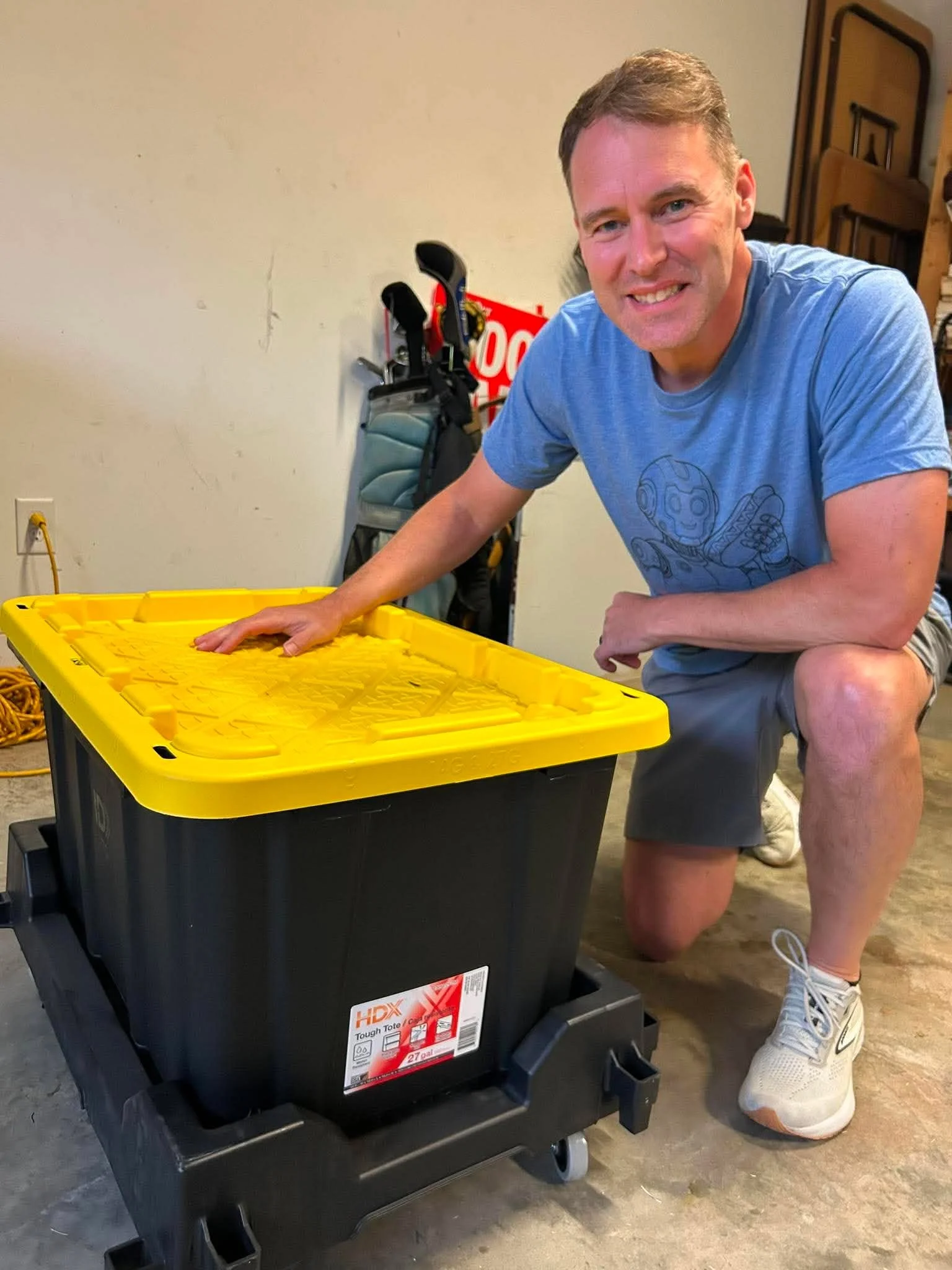 A man kneeling beside a large black and yellow trash bin, smiling at the camera, in an indoor space with a white wall, golf clubs, and folded tables in the background.