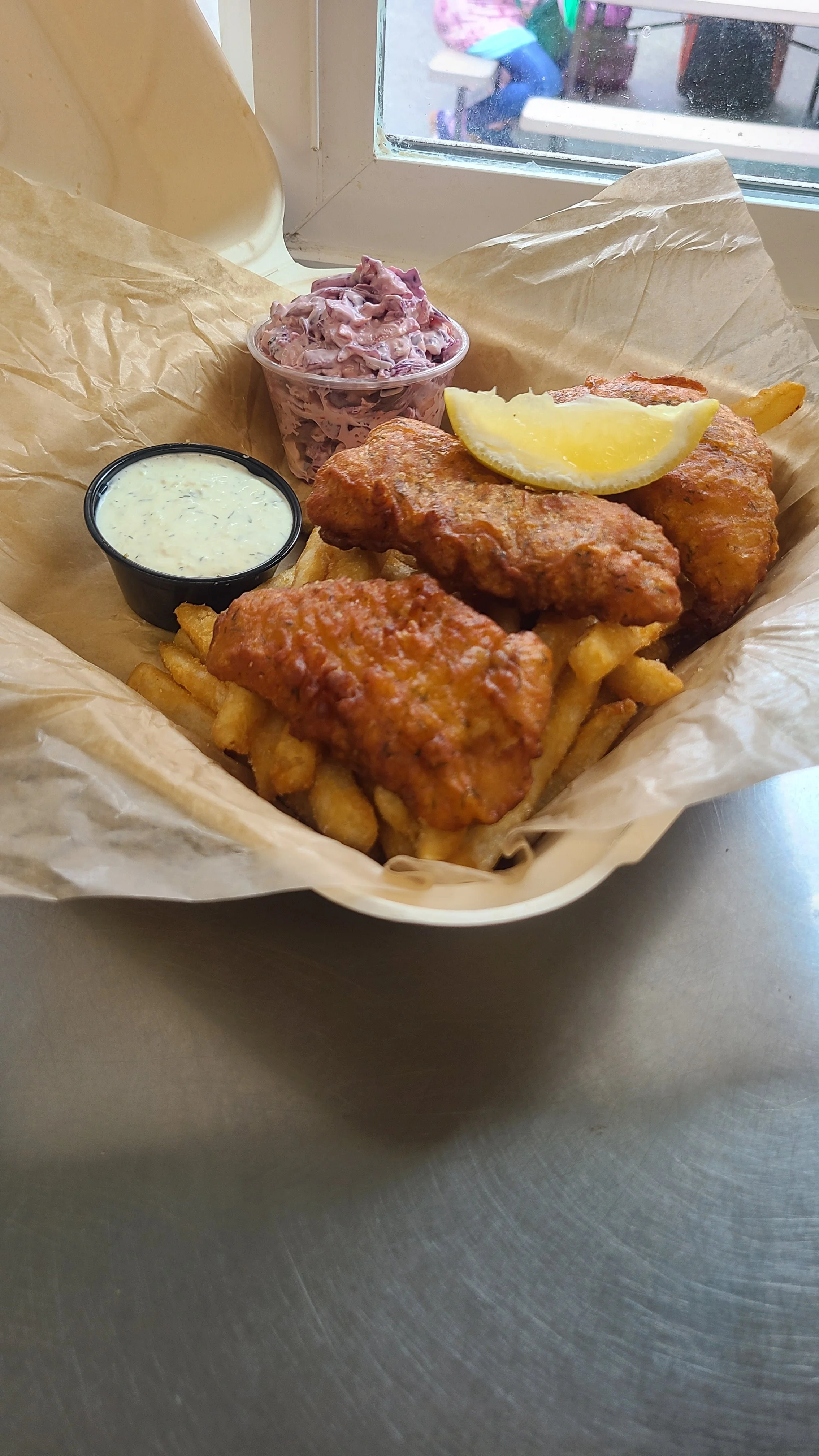 Fried fish fillets served on a bed of French fries, garnished with a lemon wedge, with a side of coleslaw and tartar sauce, in a cafeteria-style tray near a window.