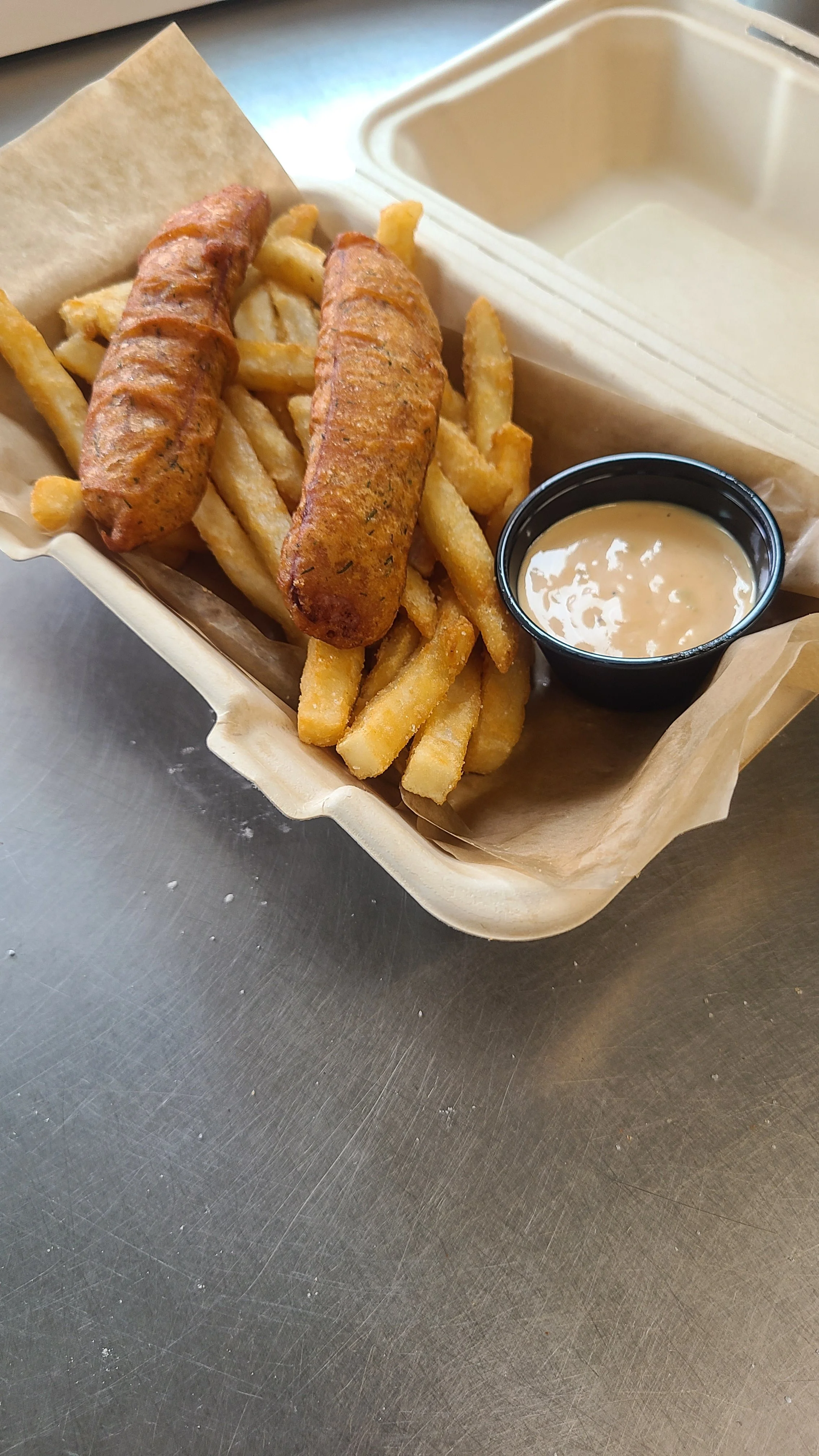 French fries served with two grilled sausages and dipping sauce in a takeout container.
