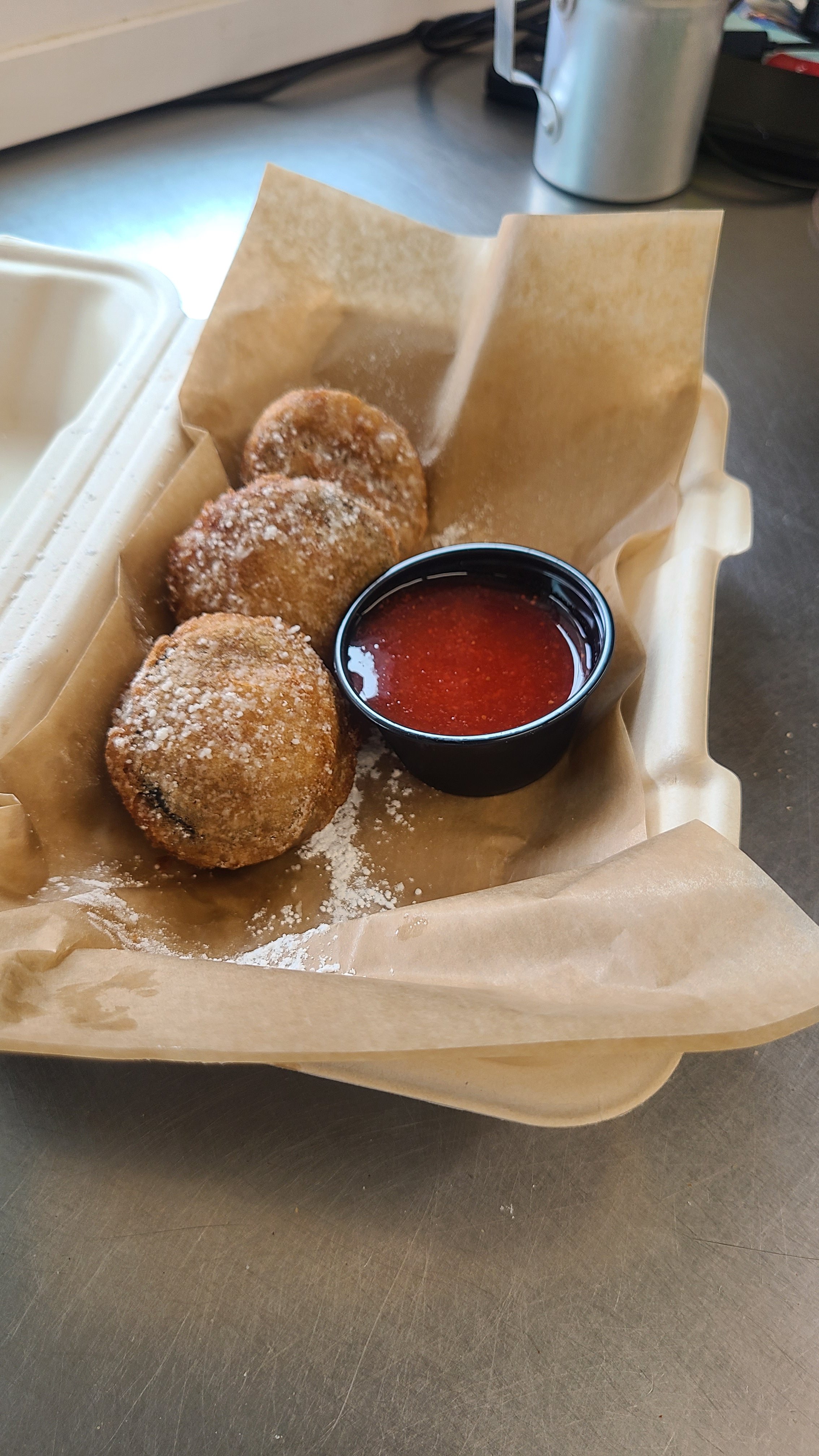 Three fried mozzarella sticks covered in powdered sugar, served with a small cup of red dipping sauce in a paper-lined basket.