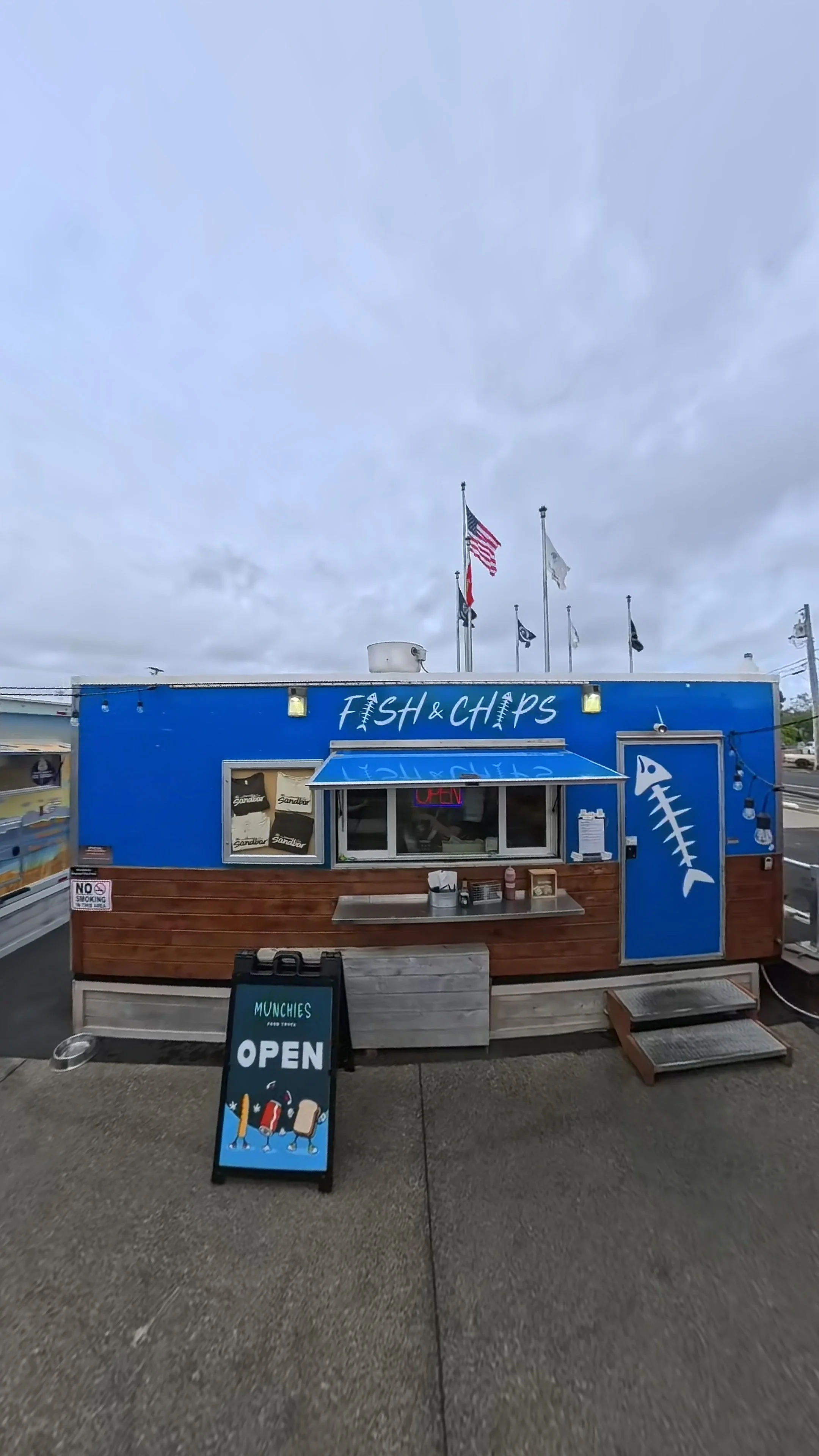 Blue food truck named 'Fish & Chips' with an illustration of a fish skeleton, serving food, with flags and an American flag in the background, and a sign indicating it is open.