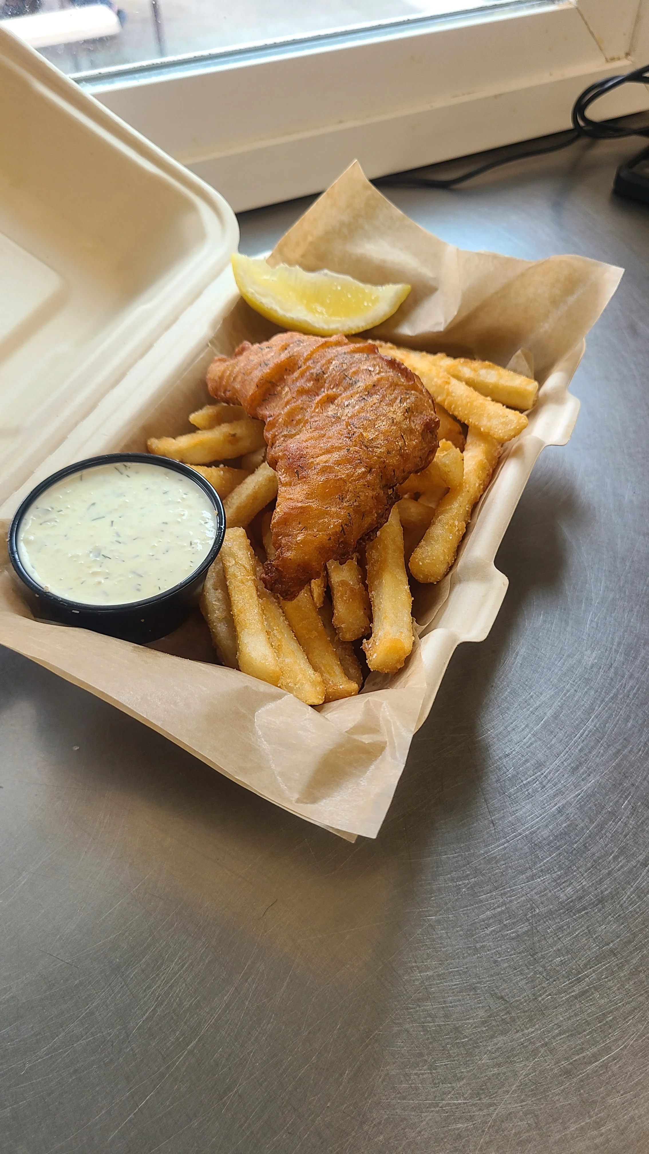 Battered fried fish fillet with French fries, lemon wedge, tartar sauce in a small container, on a paper-lined takeout box.
