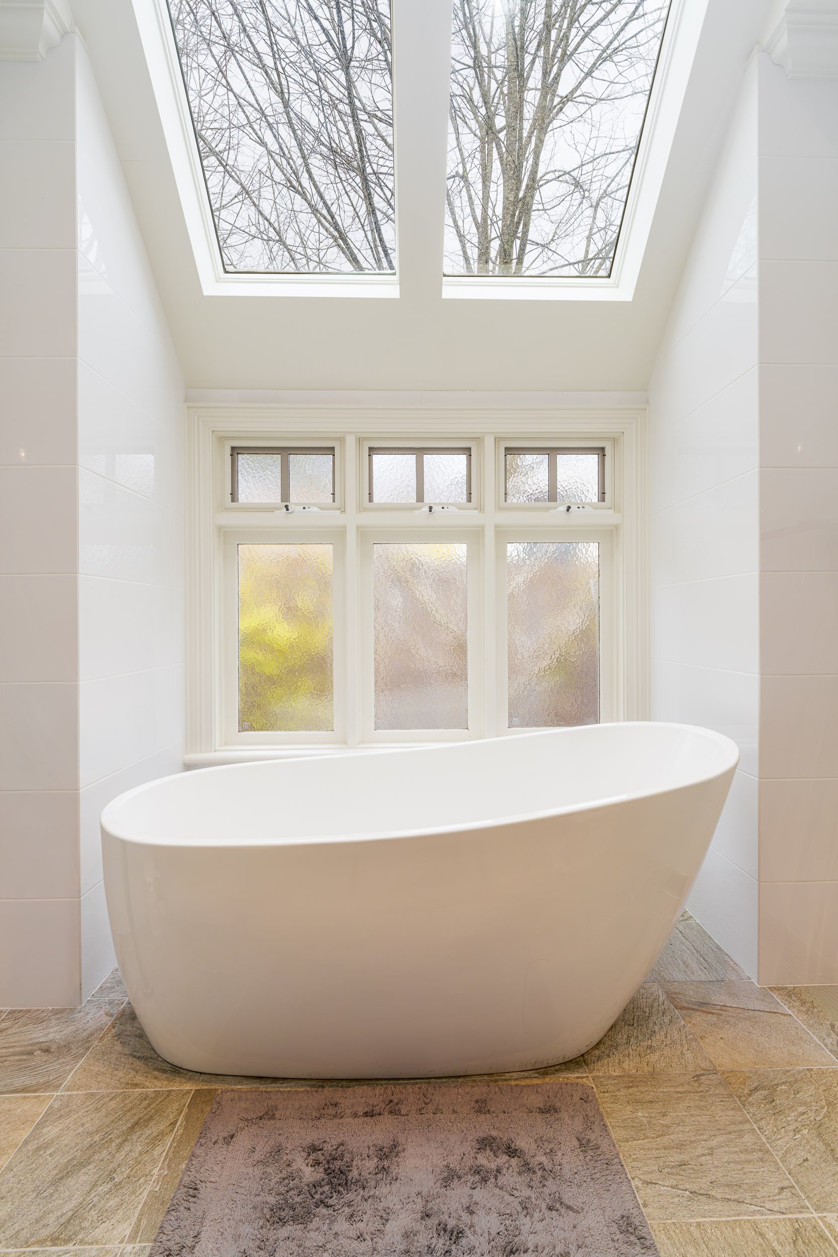 A modern bathroom with a large free-standing white bathtub placed in front of a window with frosted glass, above a beige and gray cloud-patterned rug. The ceiling has a skylight with a view of tree branches.