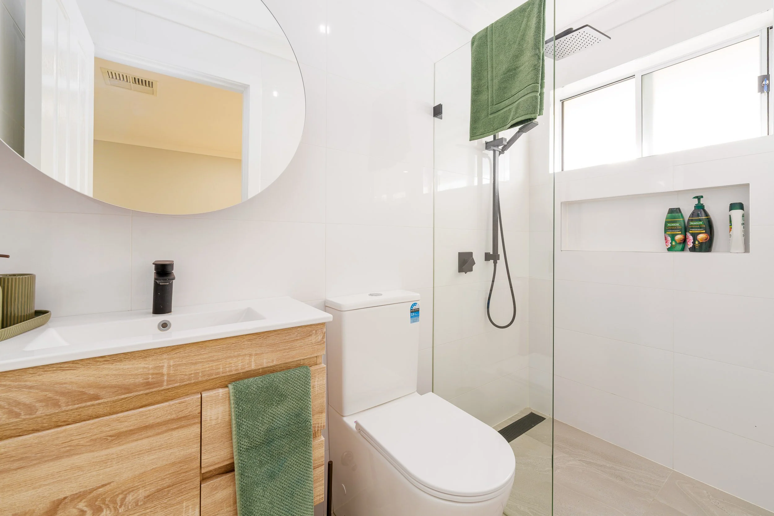 Modern bathroom with white walls, wooden vanity, oval mirror, green towel, and glass shower enclosure. Shower has a rainfall showerhead and built-in shelf with toiletries. Natural light coming through a window.