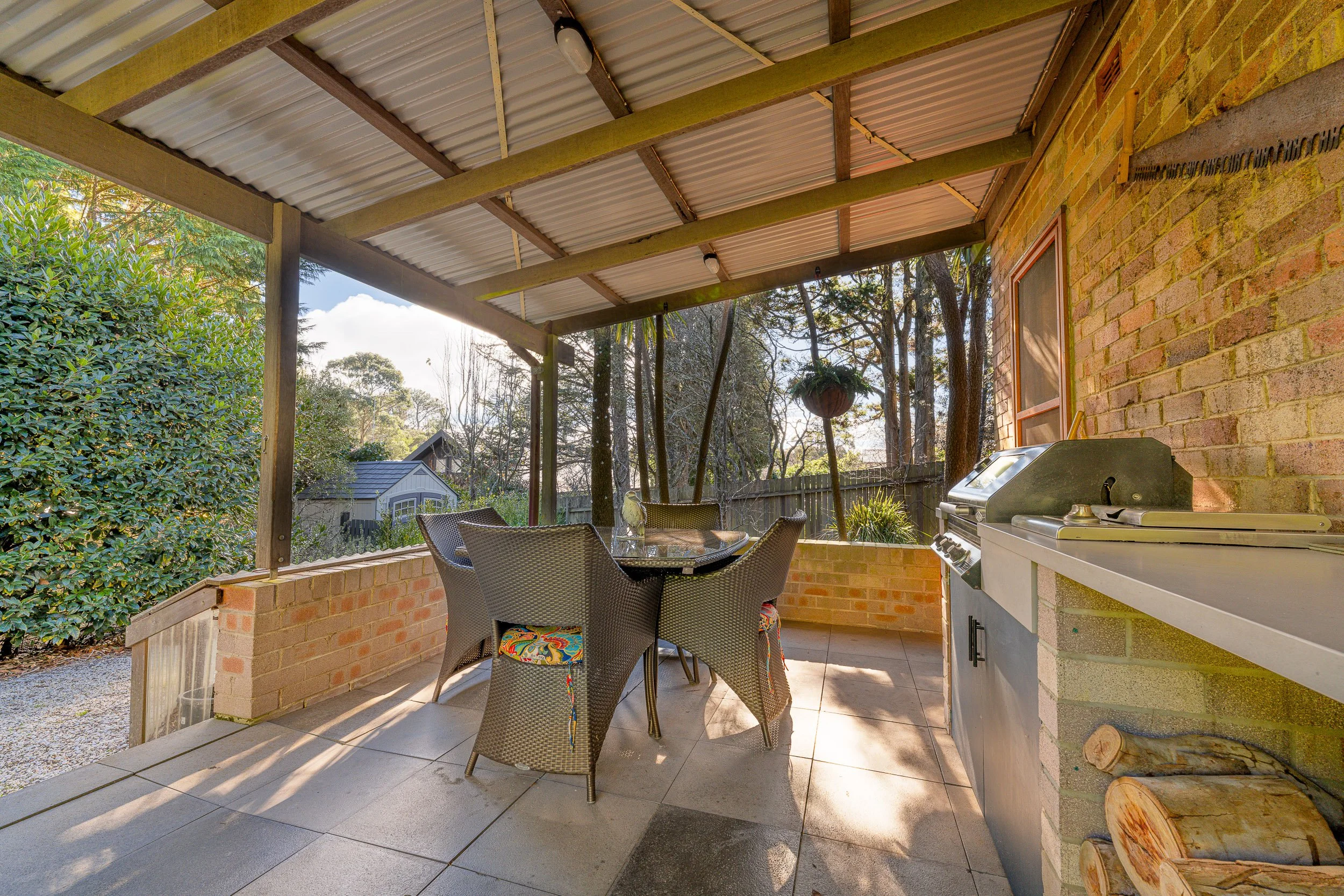 Covered outdoor patio with a brick wall, patio table with four chairs, a grill, and surrounded by trees and greenery.