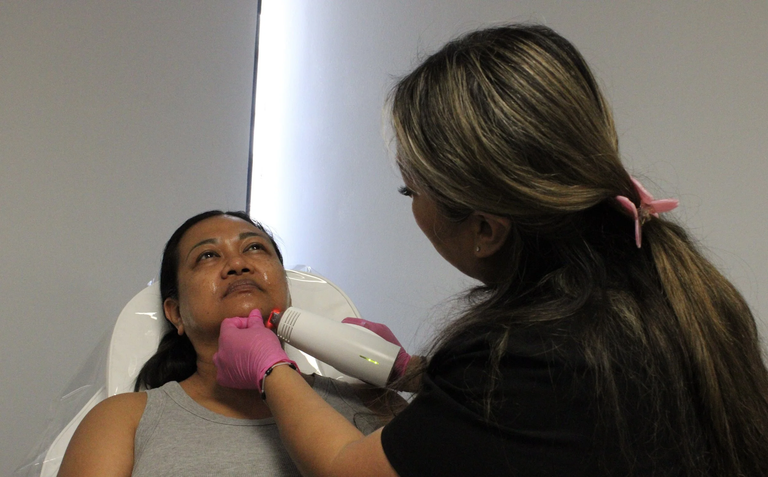 Woman lying on a medical examination table receiving laser or light therapy from a healthcare professional in a clinical setting.