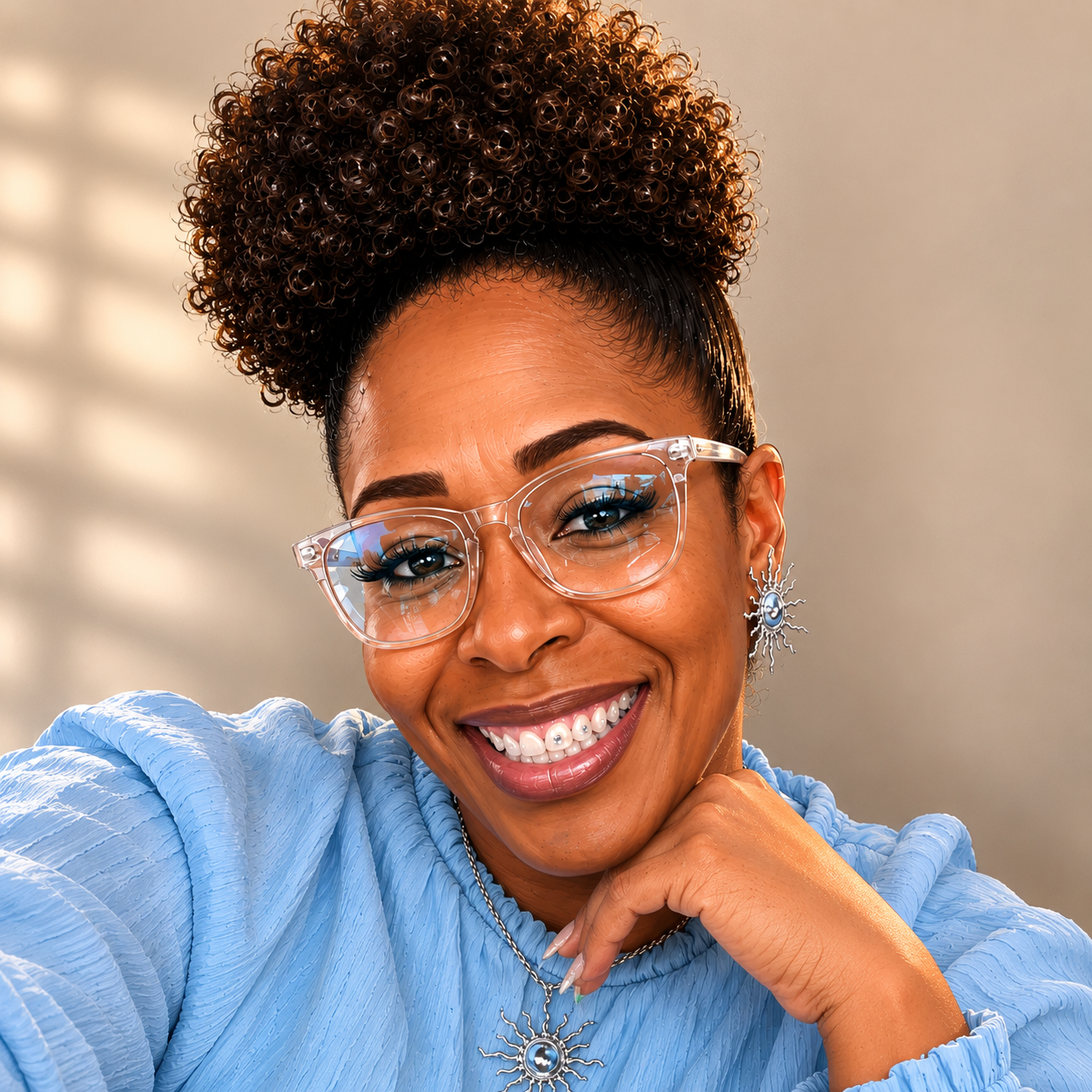 A smiling woman with curly hair, wearing clear glasses, silver sun-shaped earrings, and a blue textured top.