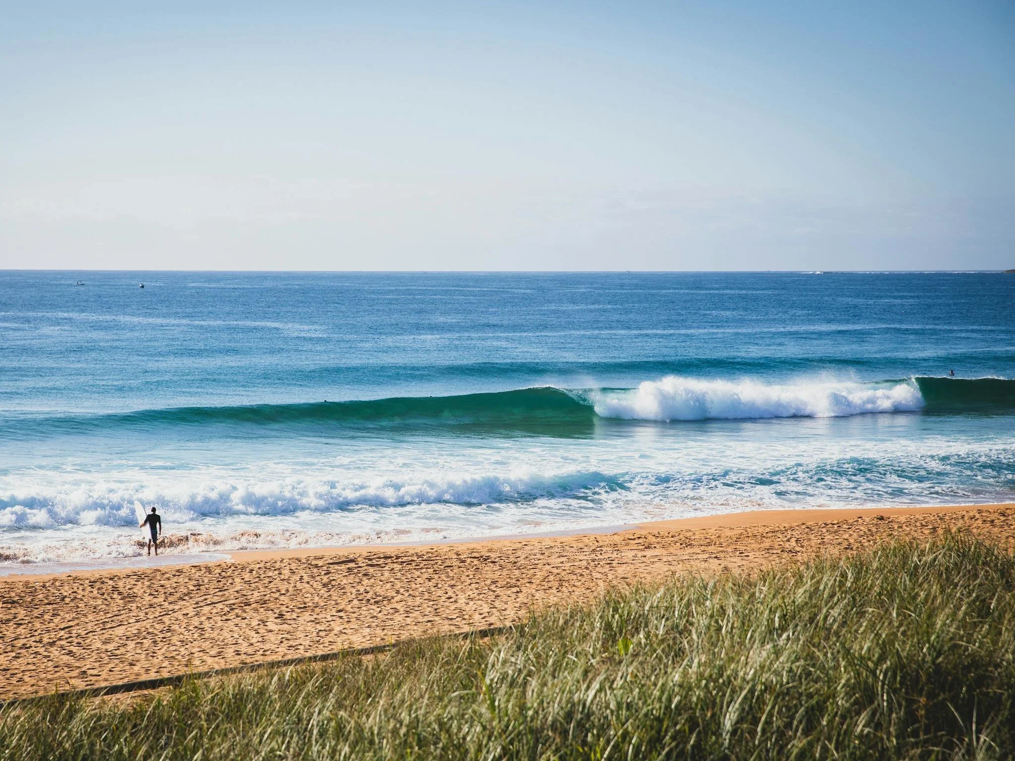 A person walking along the shoreline with a surfboard, the ocean with waves, and a blue sky in the background.