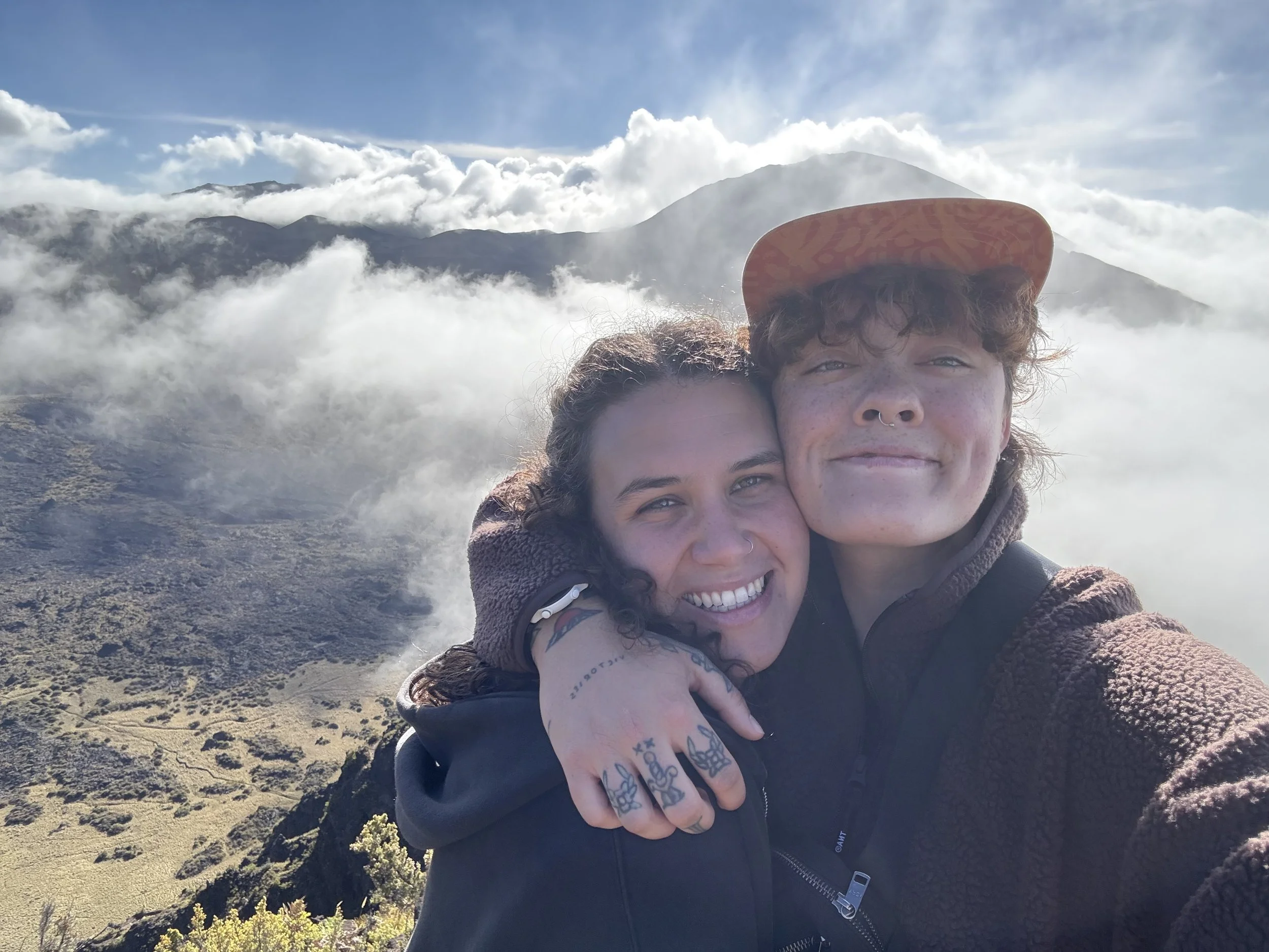 Two people embracing on a mountain with clouds and distant mountains in the background.