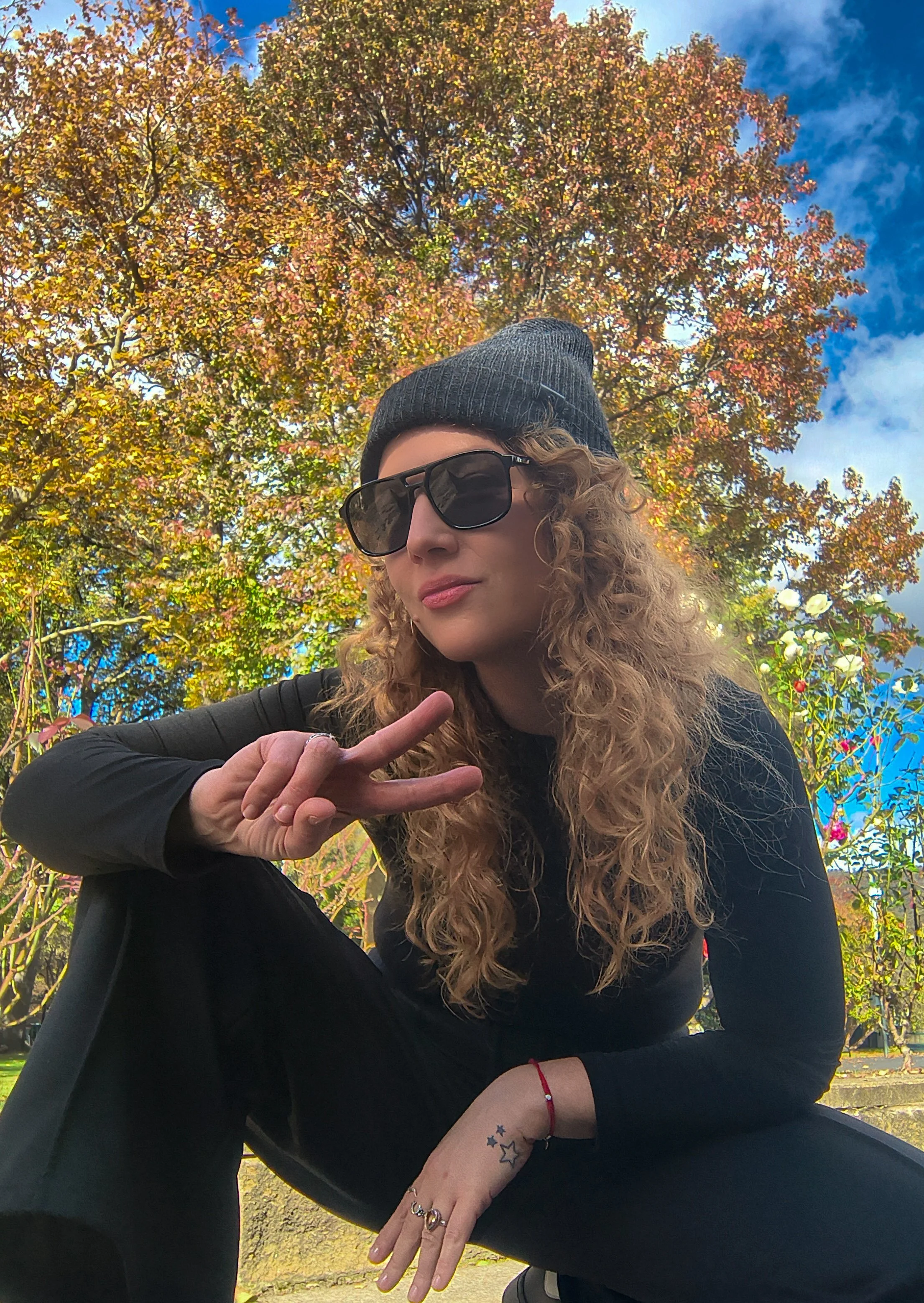 woman showing the peace sign with her fingers, half kneeling, wearing black in  a park with a large tree in the background