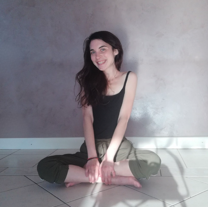 Maëva assise en tailleur au sol, posture de méditation, souriant à la caméra