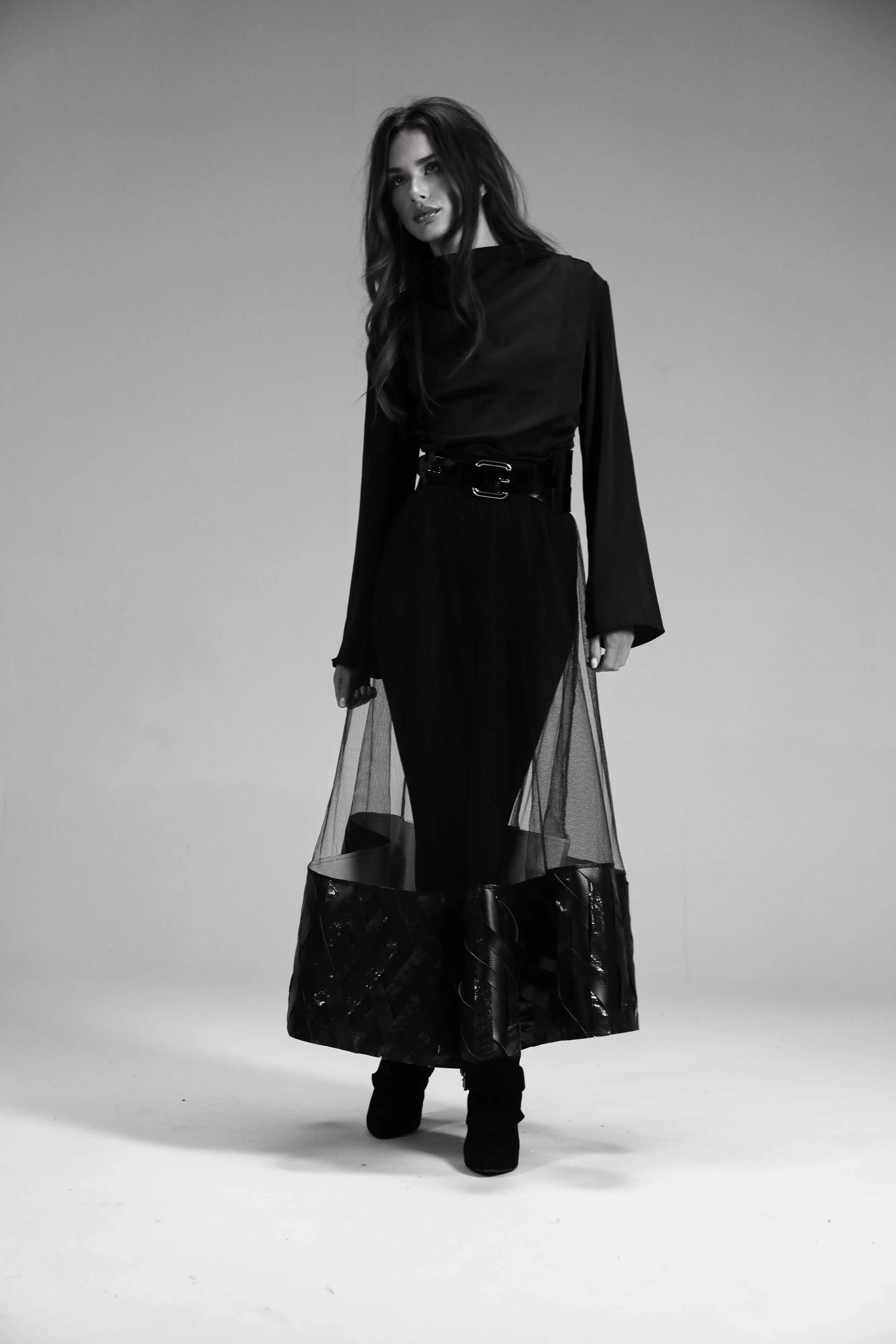 A woman with long hair, wearing a black long-sleeved top and a sheer layered skirt, standing in a studio with a plain background, facing the camera.