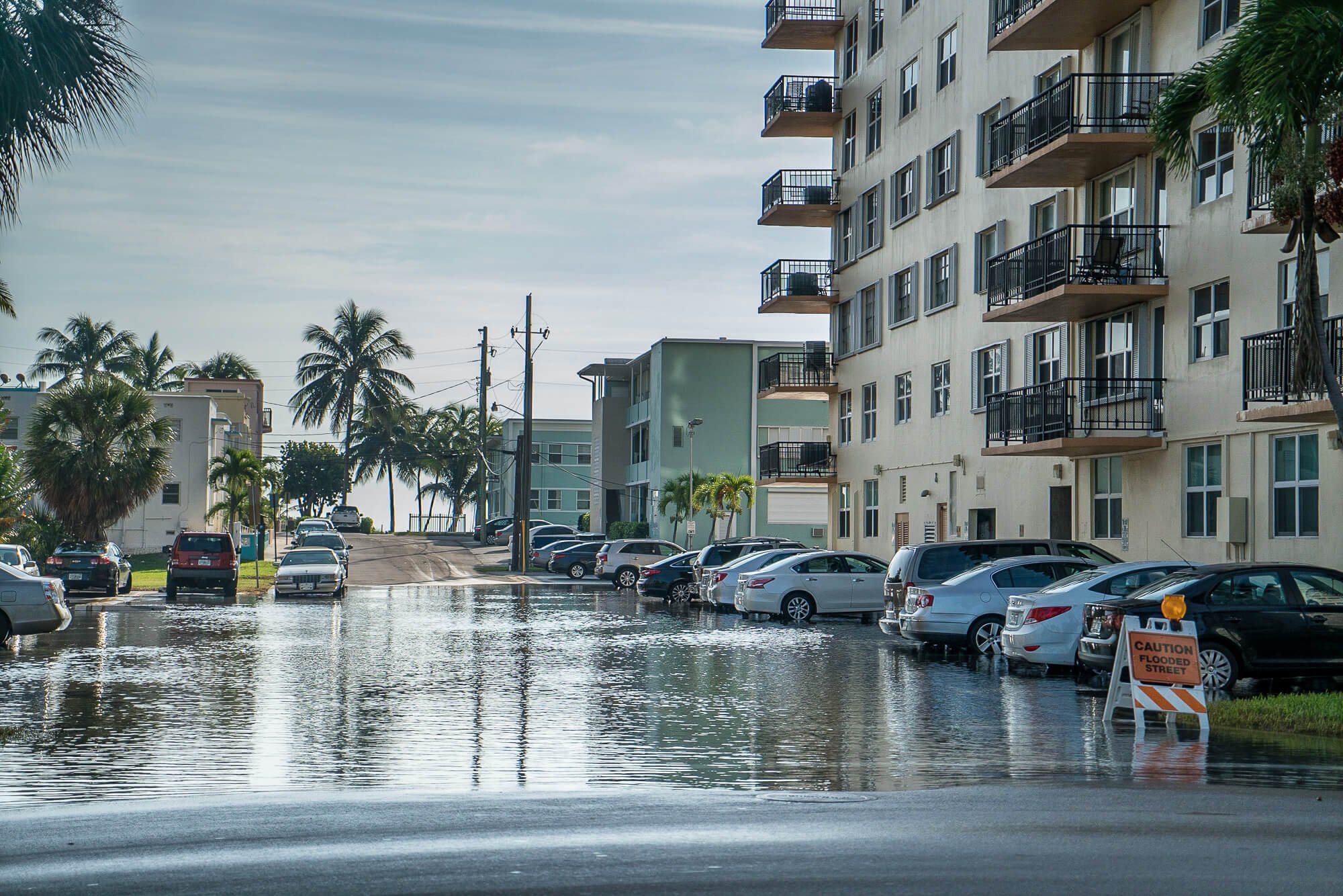 Flooded street in Miami-Dade with a "Caution Flooded Street" sign and parked cars surrounded by standing water